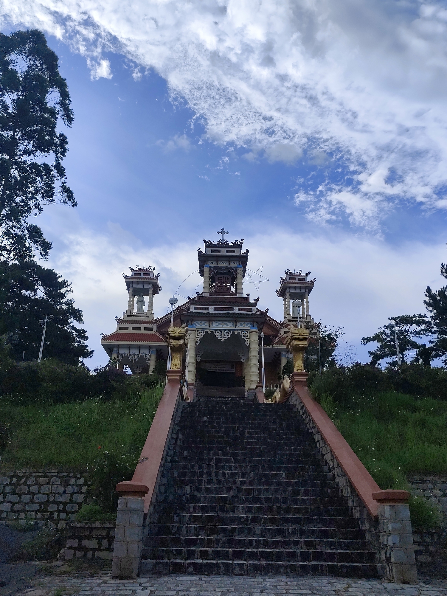 The image shows the Chùa Linh Sơn temple in Cam Ly - Đà Lạt, Vietnam. The temple is atop a hill accessed by a long flight of stone steps with pink/beige-colored railings. The temple structure consists of a central covered area flanked by two smaller tower-like structures. Each tower contains a statue. A cross adorns the top of the central structure. The sky is blue with scattered white clouds. Trees are visible on either side of the steps and around the temple. The grass on the hill is green.
