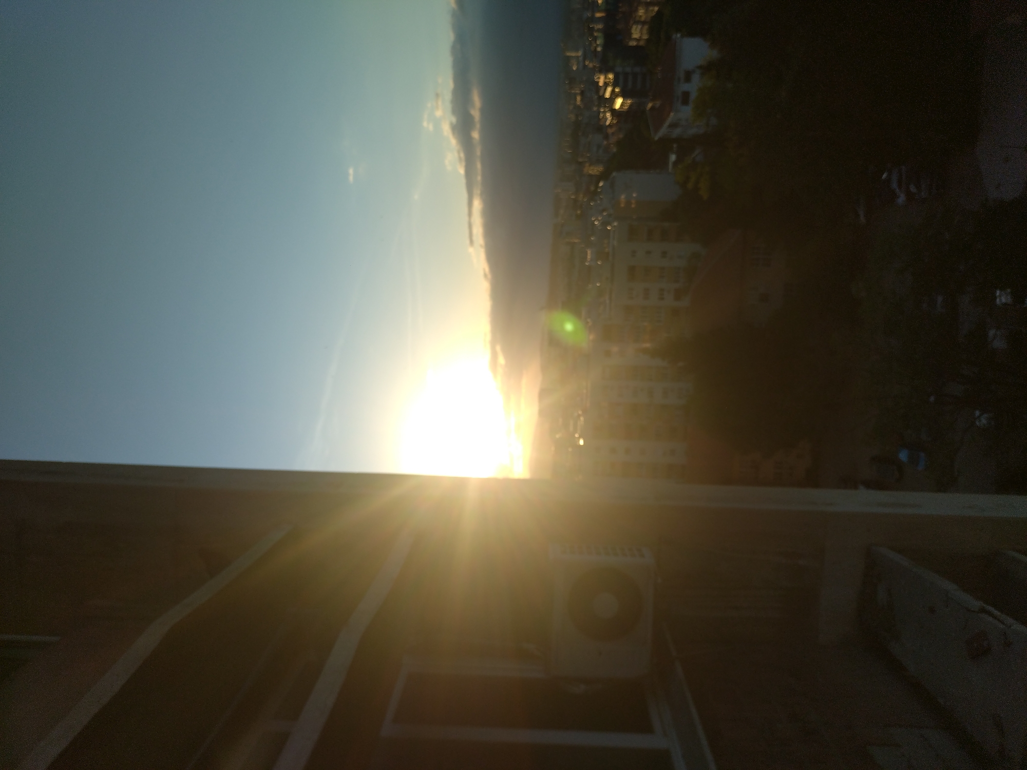 The image shows a cityscape at sunset, likely in Stara Zagora, Bulgaria, based on the cityscape view. The sun is low on the horizon, creating a bright glare. The sky is blue with some cloud cover. In the foreground, there is a rooftop with an air conditioning unit and a window. Trees and building details can be seen in the midground.