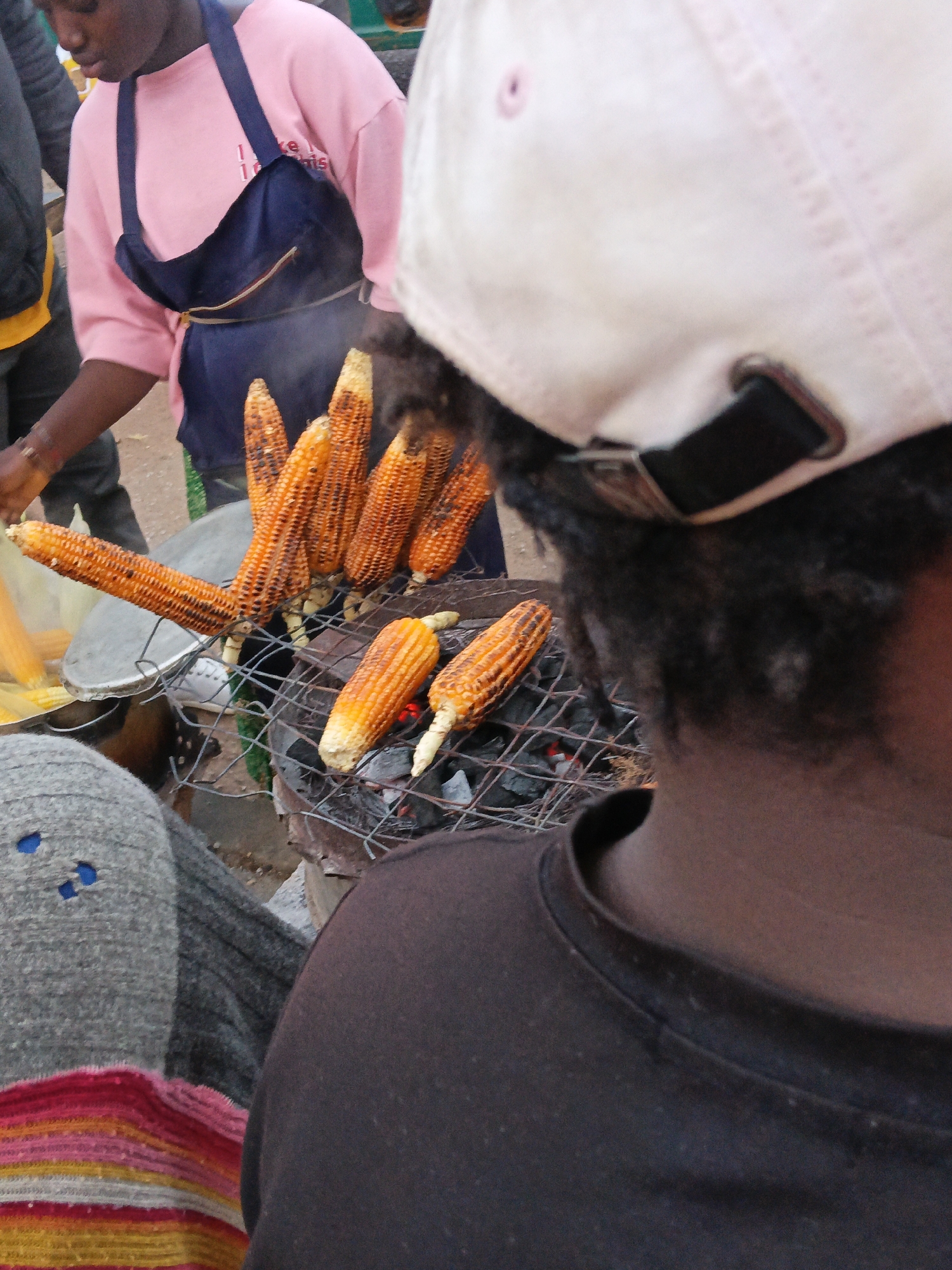 The image shows a close-up view of a street food vendor grilling corn on a charcoal grill. In the foreground, the back of a person wearing a dark brown t-shirt and a light-colored baseball cap is visible. The vendor, likely a woman based on the pink t-shirt and blue apron, is to the left, actively managing the corn. Several ears of corn are arranged on a wire rack over glowing red coals. The corn is charred in places, indicating it is being roasted. Behind the vendor, a large metal pot or basin is visible. The scene appears to be outdoors, with other people and a blurred background suggesting a busy street or market environment. The ground is paved. The overall impression is one of a casual outdoor food preparation setting.