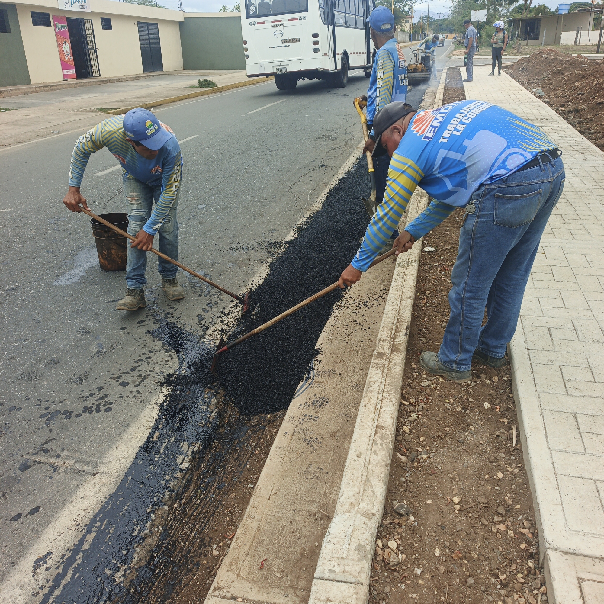 The image captures road maintenance work in the city of Tinaquillo, Venezuela. Three workers are seen patching a section of the asphalt road with dark material. One worker in the foreground spreads the material with a rake, while another uses a similar tool and a bucket. A third worker is seen using a shovel on the asphalt. All workers wear blue shirts and hats with a logo. A white bus is stopped on the road, and other workers are present in the background, along with construction equipment. A newly paved section of a walkway runs adjacent to the road. The scene includes asphalt, concrete edges, and a dirt area between the road and the walkway.