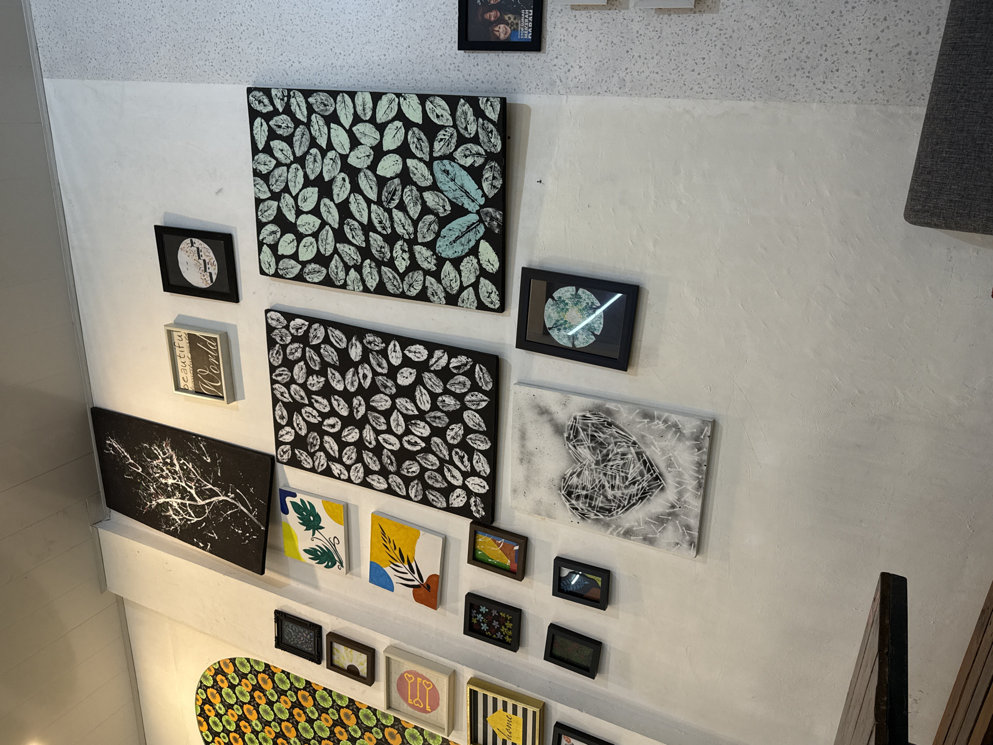 The image shows a wall decorated with a collection of framed artworks of various sizes and styles. The artworks include paintings and prints featuring abstract designs, botanical motifs, and geometric patterns. The wall has a slightly textured surface and appears to be painted in a light color, possibly white or off-white. The location of the scene is implied to be in Lhokseumawe, Indonesia.