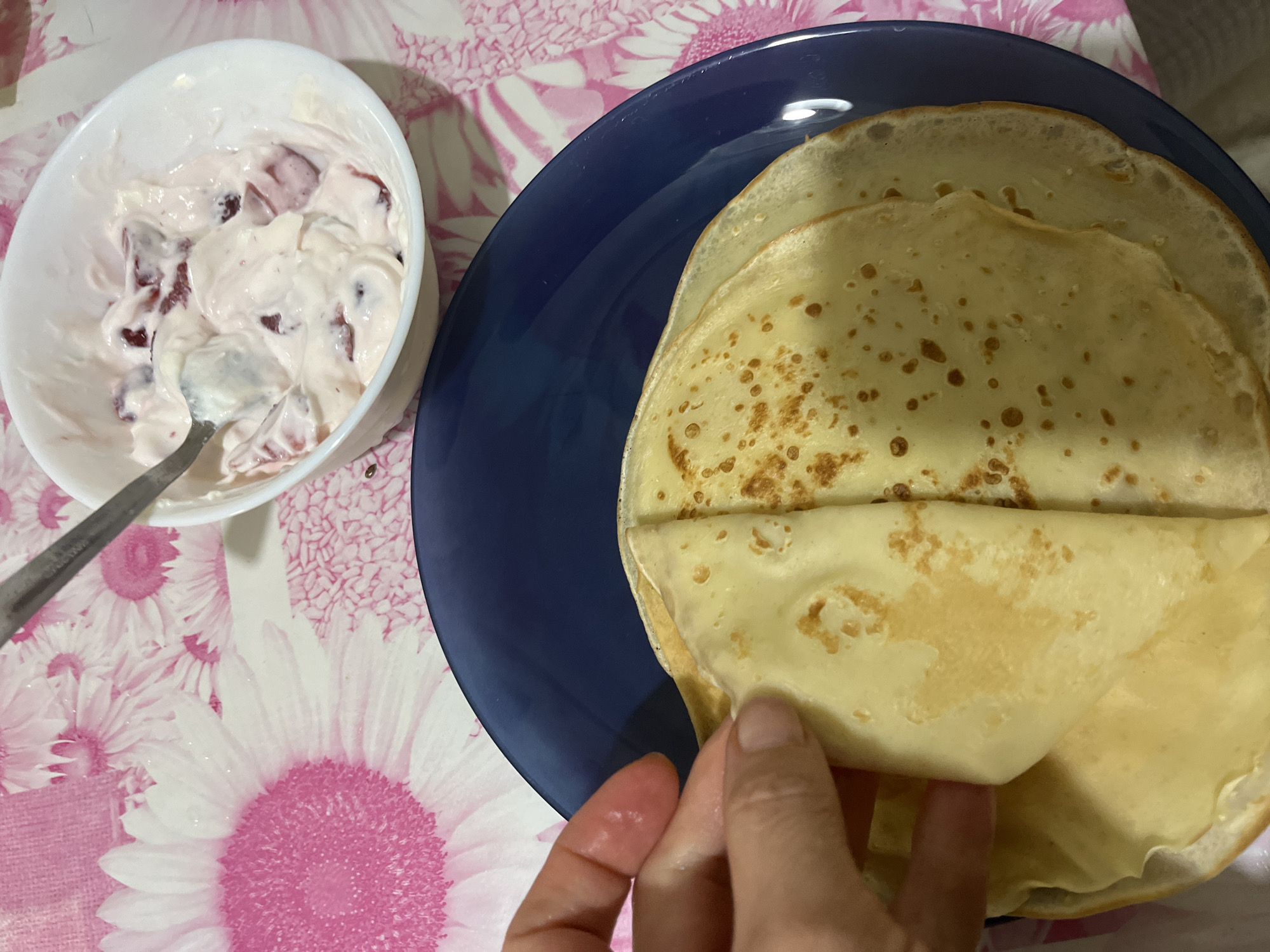 A close-up shot features a plate of crepes and a bowl of what appears to be yogurt with fruit on a patterned tablecloth. A hand is in the process of folding one of the crepes on the blue plate. The bowl of yogurt is white and has a silver spoon in it. The tablecloth is white with a pink floral pattern. The location is presumed to be in Alanya, Turkey.
