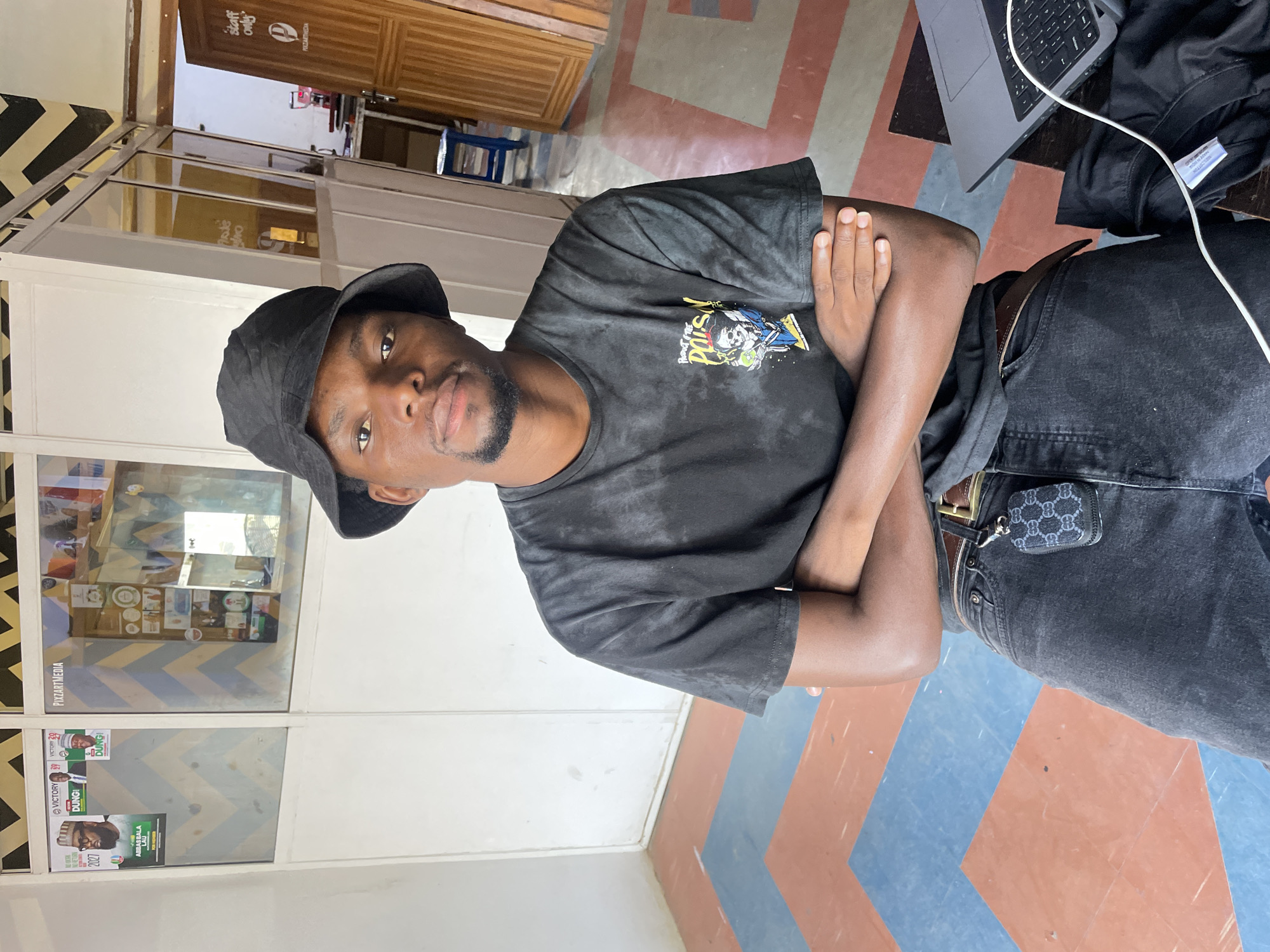 The image, taken in Jos, Nigeria, features a young Black man posing indoors, looking directly at the camera with a neutral expression. He wears a black bucket hat, a dark tie-dye t-shirt with a colorful graphic design, dark jeans, and a brown belt with a small, patterned pouch clipped on. His arms are crossed over his chest.
The setting appears to be a brightly lit commercial or office space. The floor has a distinctive geometric pattern of reddish-brown and blue-gray stripes. A large glass partition dominates the background to the man's left, reflecting elements and displaying several political campaign posters. To his right, an open laptop and a white charging cable rest on a dark surface. A wooden door in the background bears the text "STAFF ONLY" and "PIXZARTMEDIA."
The activity depicted is a casual pose for a photograph. Visible text includes "STAFF ONLY," "PIXZARTMEDIA" (also seen as "PULZARTMEDIA" in reflection), "NO RETURN" (reflected), and slogans on campaign posters such as "VICTORY," "DUNG!", "ABBAS BALA LAU," and "2027." Reflections also show "PRODUCT & THE." The presence of political campaign materials and the business name suggest a local media or creative office environment.