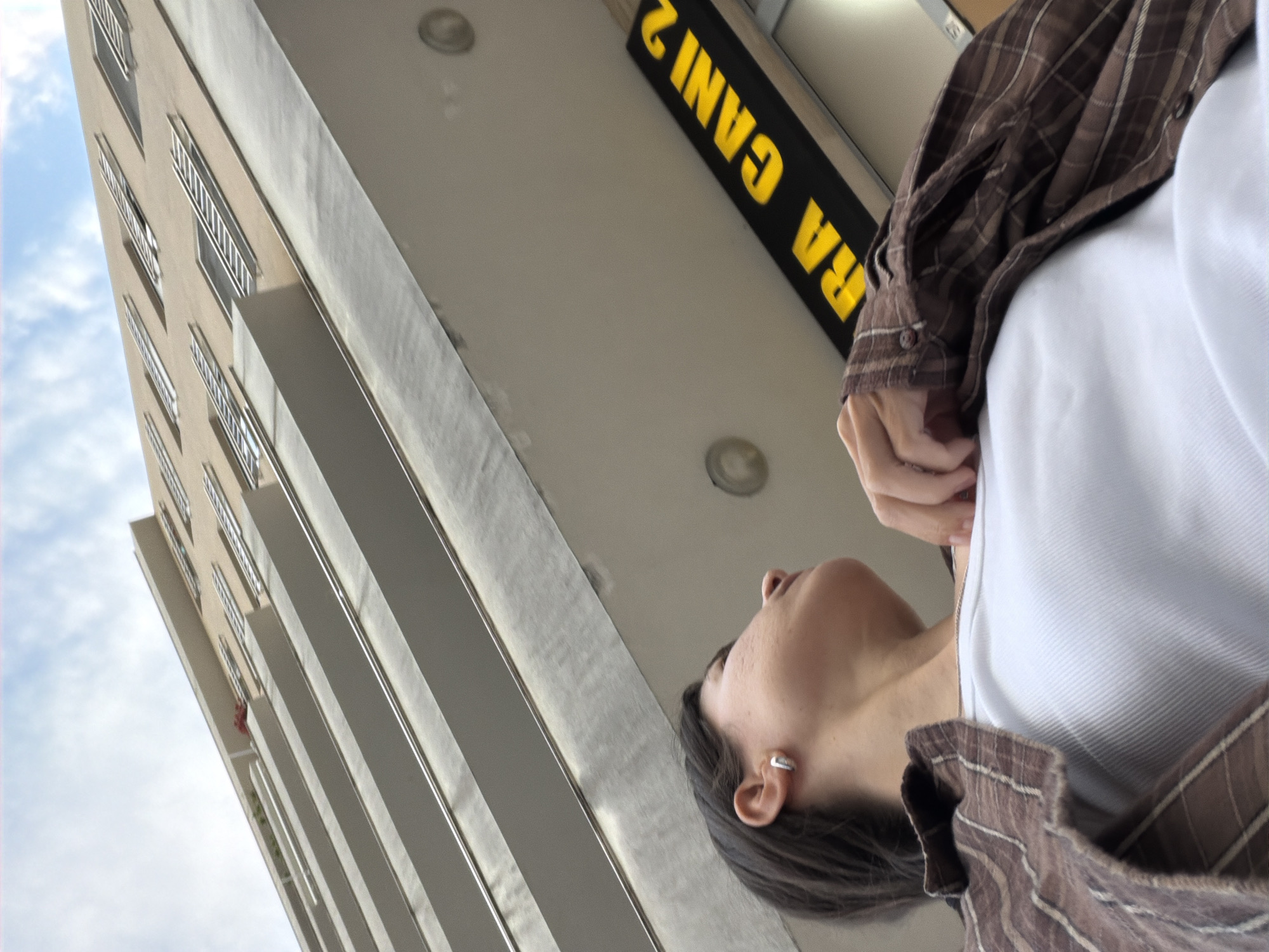 An upward-angled shot captures a partial view of a person alongside a modern building in Budva, Montenegro. The composition is dynamic, with the horizon tilted diagonally.
On the right side of the frame, the upper torso and head of a person are visible in profile, looking upwards. They have dark hair pulled back, a visible ear adorned with a simple silver hoop earring, and fair skin with some texture. They are wearing a white undershirt beneath an unbuttoned brown plaid or striped long-sleeved shirt. Their hands are partially visible near their chest, suggesting a relaxed posture.
The left and upper-middle sections of the image are dominated by the facade of a multi-storied building. It is light-colored, possibly beige or light gray, with numerous rectangular windows and balconies featuring white, horizontally barred railings. The building appears to be part of an urban complex.
Above the person and beneath an overhang or ceiling, a black rectangular sign with bright yellow text is prominently displayed. Due to the camera's low angle and the sign's orientation, the text appears reversed and upside down. When corrected for perspective, the visible text reads "RA CANI 2".
The sky, visible in the upper left, is bright blue with scattered white clouds, indicating daytime. The overall scene suggests a casual moment of observation or passage through an urban environment.