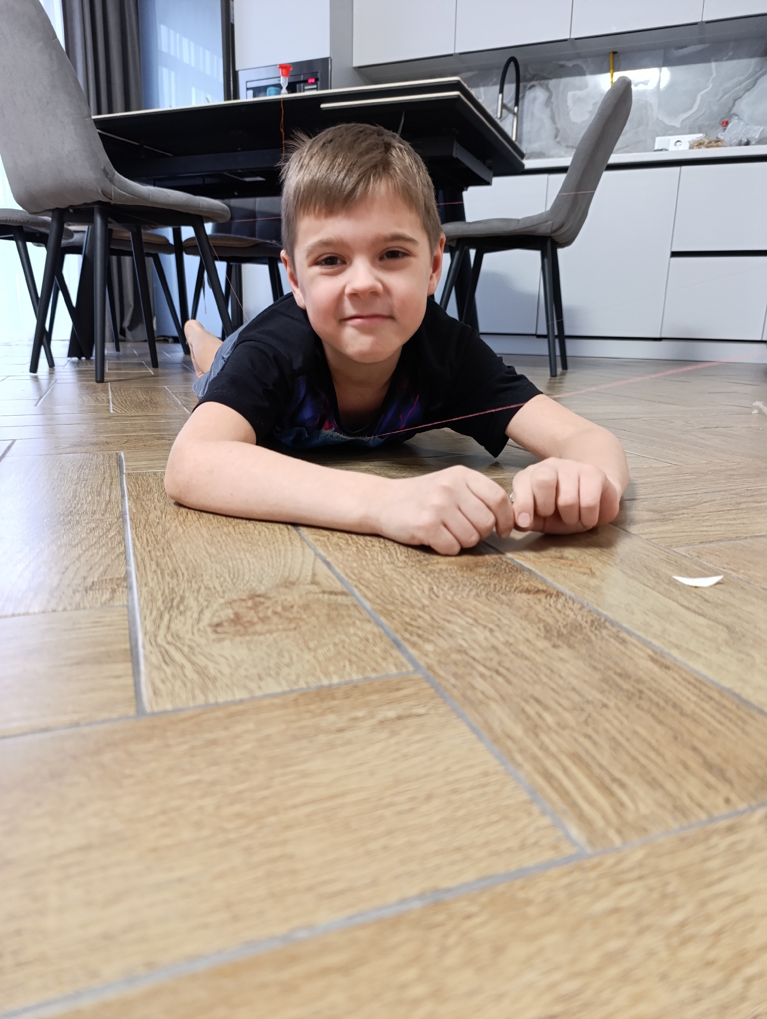 A young boy lies prone on a light brown wooden floor, possibly laminate or wood-look tile, with light-colored grout lines. He faces the camera with a slight smile. He is wearing a black short-sleeved shirt. A thin red string runs taut across his upper torso and hands. A small, white object, possibly a feather or small piece of paper, sits on the floor near his hands.
In the background, there is a black table with chairs, and a kitchen counter with white cabinets and a marbled backsplash. A microwave and a faucet are visible on the counter.
The photograph appears to be taken indoors, possibly in a home or apartment located in Chaikovsky, Russia. The location cannot be verified.