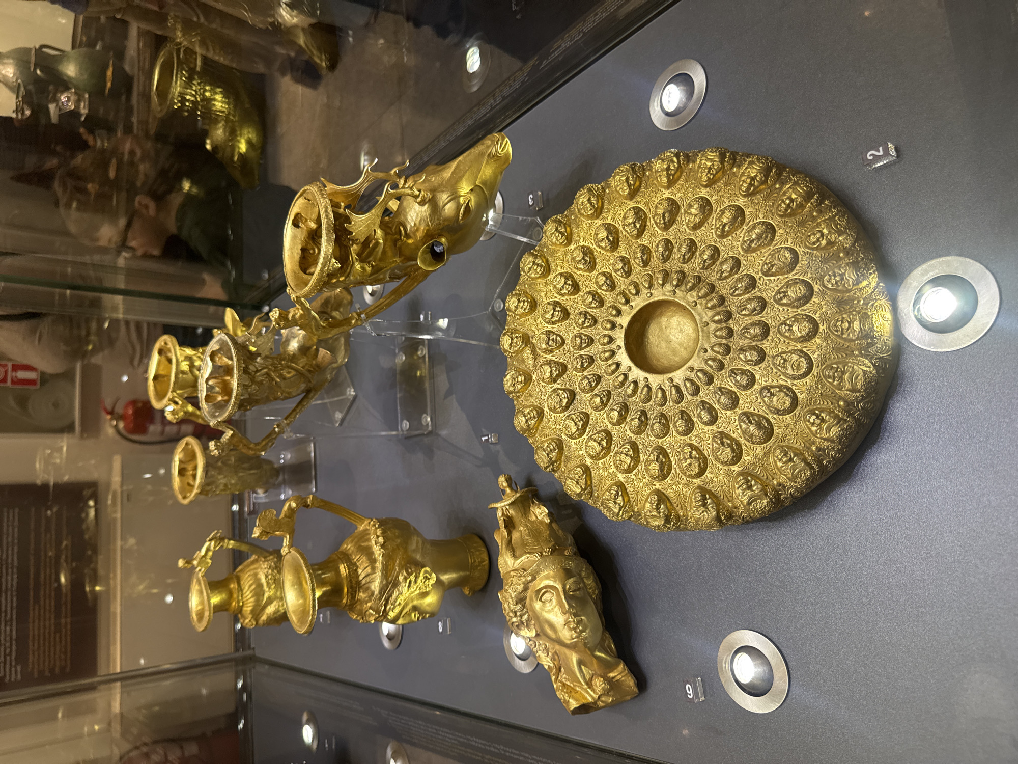 The image shows a display of gold artifacts within a glass case, likely in a museum in Plovdiv, Bulgaria. The artifacts include several rhytons (drinking vessels) shaped like animal heads or bodies, a shallow dish decorated with human faces, and other ornate gold vessels. These items are arranged on a dark gray surface, seemingly inside a display case with recessed lights illuminating them. Numbers are visible next to some of the artifacts, presumably for identification or cataloging purposes. Reflections of the museum visitors and the surrounding environment can be seen on the glass of the display case.