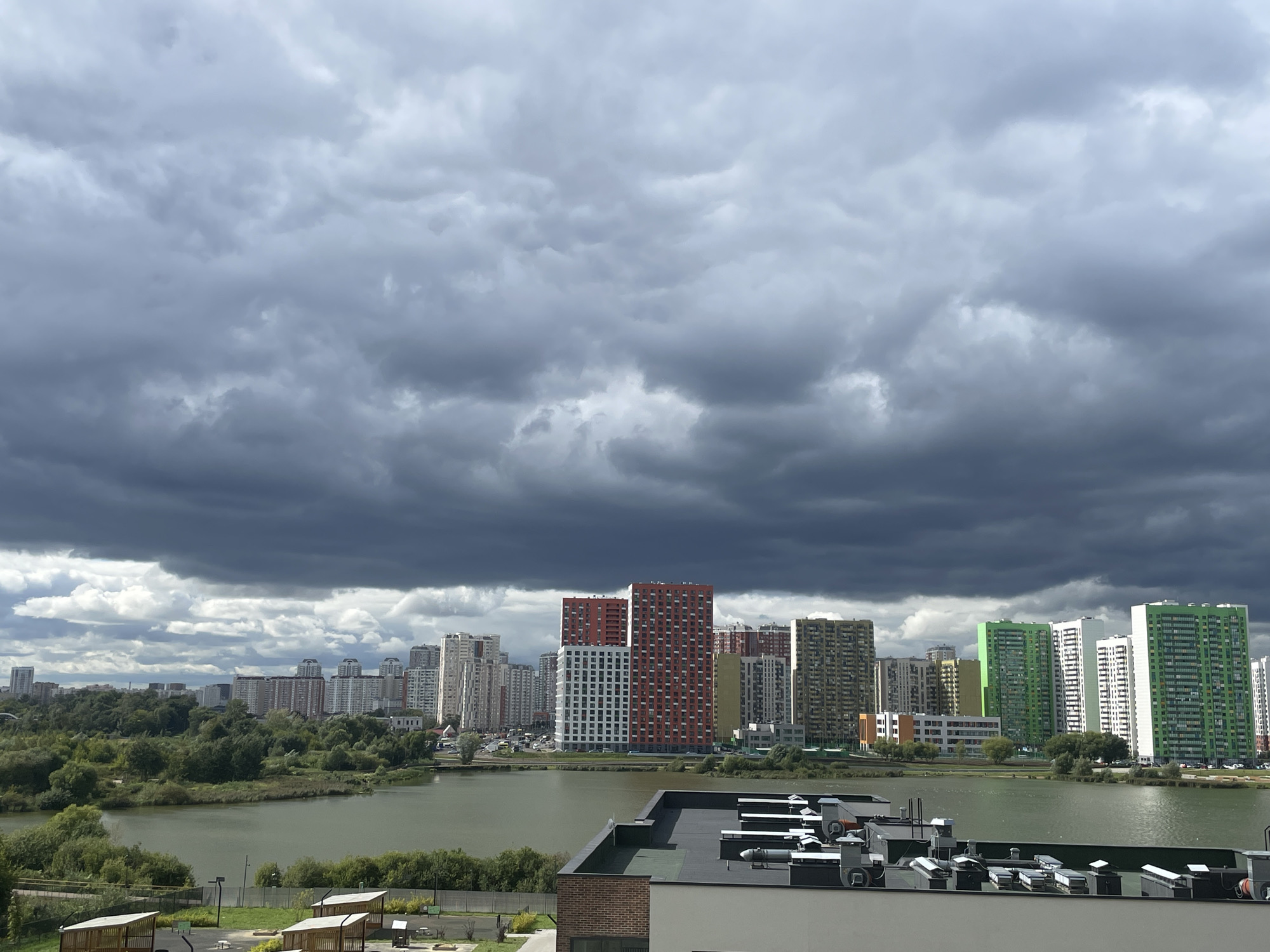 The image captures a view of a lake with a city skyline in the background under a dramatic, overcast sky filled with dark, heavy clouds. In the foreground, the rooftop of a modern building is visible, featuring various ventilation units and HVAC equipment. Beyond the lake, numerous high-rise apartment buildings are densely packed, displaying a variety of architectural styles and colors, including prominent red, white, yellow, and green structures. A strip of lush green trees and vegetation separates the lake from the urban development. The overall atmosphere suggests a possibly impending storm.