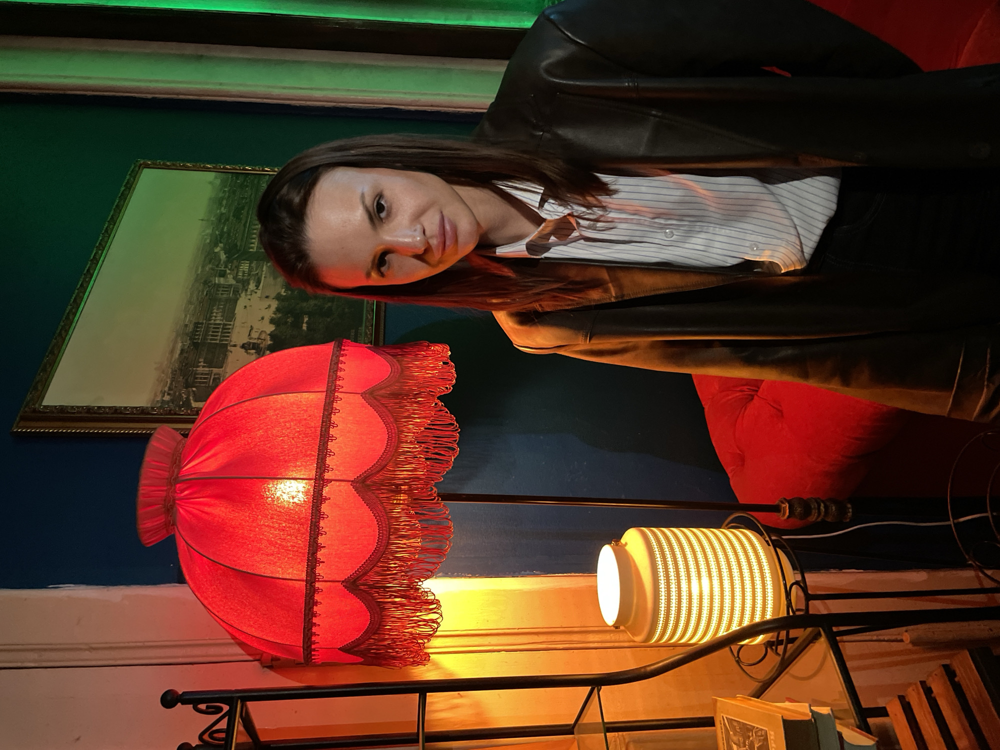 A woman is posing in front of a large red lamp with fringe. She is wearing a black leather jacket over a striped white button-down shirt and dark pants. The woman has shoulder-length brown hair and is looking at the camera with a slight smile. Behind her is a blue wall with a framed picture hanging on it. The picture appears to be a landscape of a city, potentially Saint Petersburg, Russia. To the right of the woman is a red cushion or piece of furniture. Under the red lamp, there is a second, smaller white lamp with a textured shade. Below the white lamp, some books and frames are visible on a shelf. The room has a warm, vintage atmosphere. There is also a green light visible near the top of the frame.