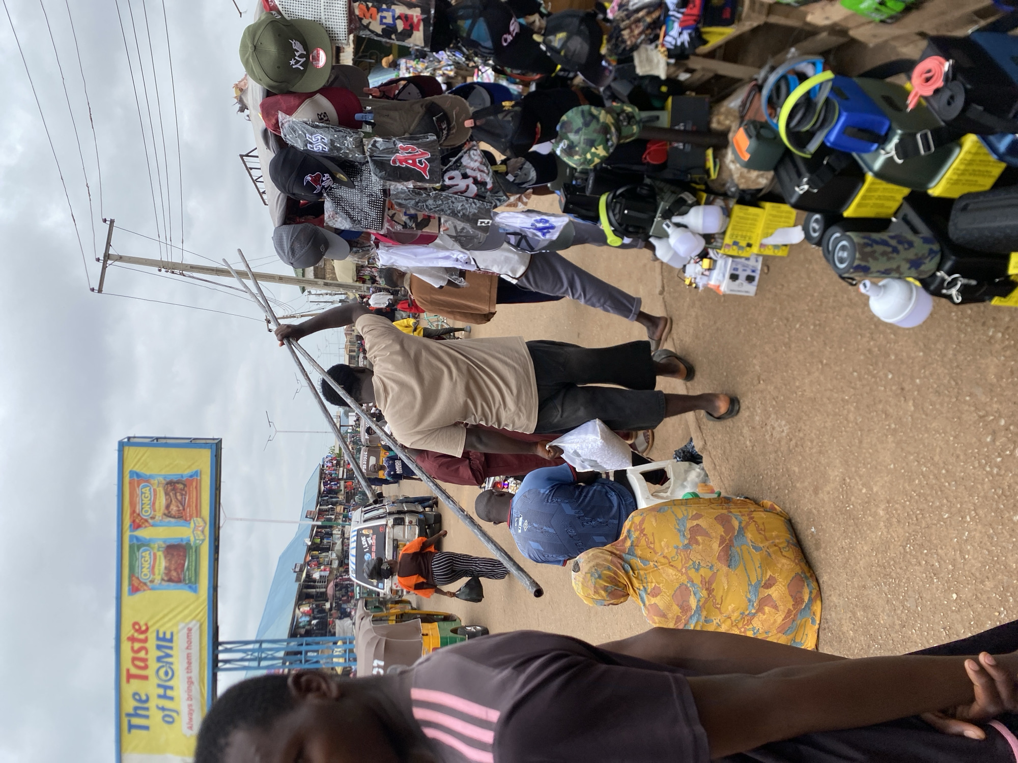 The image captures a bustling street scene in Bukuru, Nigeria. In the foreground, a person on the far left is partially visible, wearing a dark t-shirt with pink stripes. To their right, a person wearing a yellow and brown patterned headscarf and clothing is seated. Further to the right, a man in a blue t-shirt is seated on a white stool. A man in a tan t-shirt is walking to the right, carrying a white plastic bag. The background reveals a street lined with market stalls. A prominent billboard advertising "The Taste of HOME" with images of Onga seasoning is visible in the upper left. Various vehicles, including three-wheeled auto-rickshaws and a white van, are present on the street. Several market stalls are displayed, showcasing an assortment of goods such as hats, bags, and electronic items. The overall environment appears to be an outdoor market under a cloudy sky.