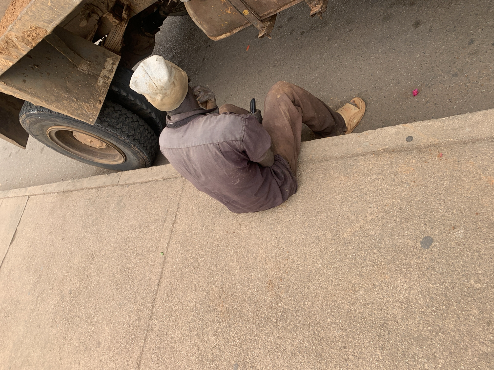 A high-angle shot depicts a man, possibly a mechanic, seated on a sidewalk adjacent to a truck, likely performing maintenance or repairs. The man wears a white hard hat, dark-colored gloves, a long-sleeved shirt, and brown pants. He holds a dark object in his gloved hands. His posture is relaxed, with his legs stretched out along the curb and his left foot clad in a sandal. The truck is partially visible in the upper left, showing a tire and rusty metal components. The surface under and around the truck is stained with dirt and oil. The sidewalk extends into the foreground, showing a sandy color and some discoloration. A fallen pink flower lies on the street next to the curb near the man's foot. The scene is typical of an urban setting in Jos, Nigeria, where trucks are often maintained on the roadside.