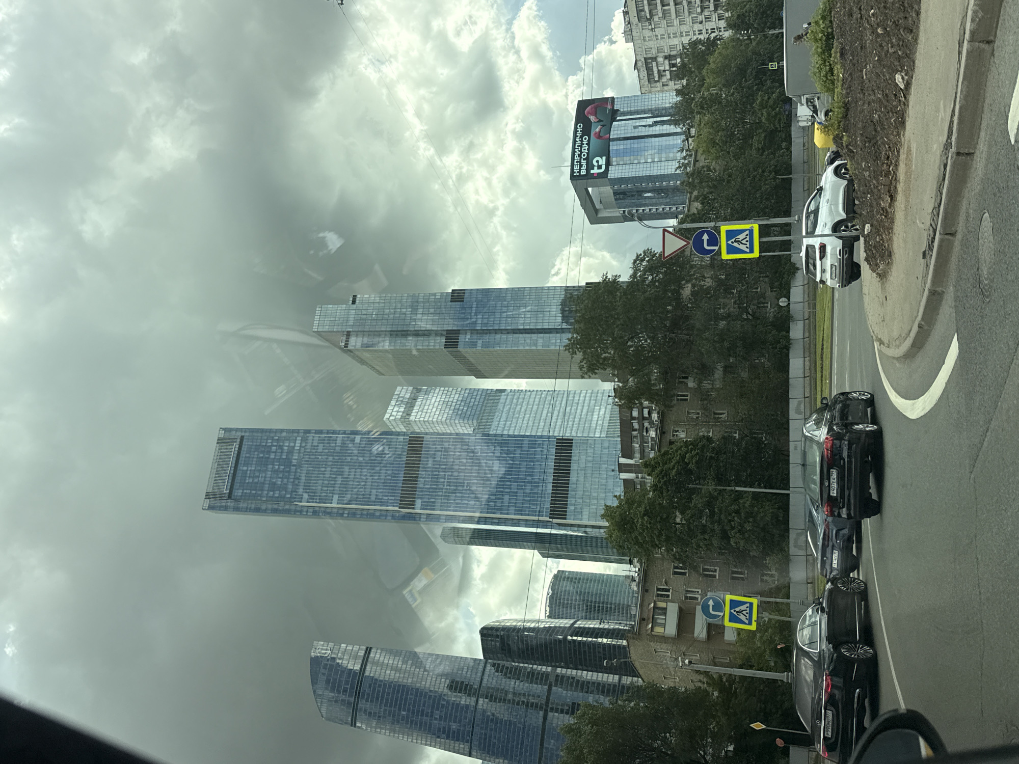 The image, taken from inside a vehicle, shows a street scene in Khimki, Russia. Several modern skyscrapers with glass facades are visible, some reflecting the cloudy sky. In the foreground, there is a road with parked black cars. Street signs indicating pedestrian crossings and turn directions are present. Buildings that are older than the sky scrappers are also in the background.