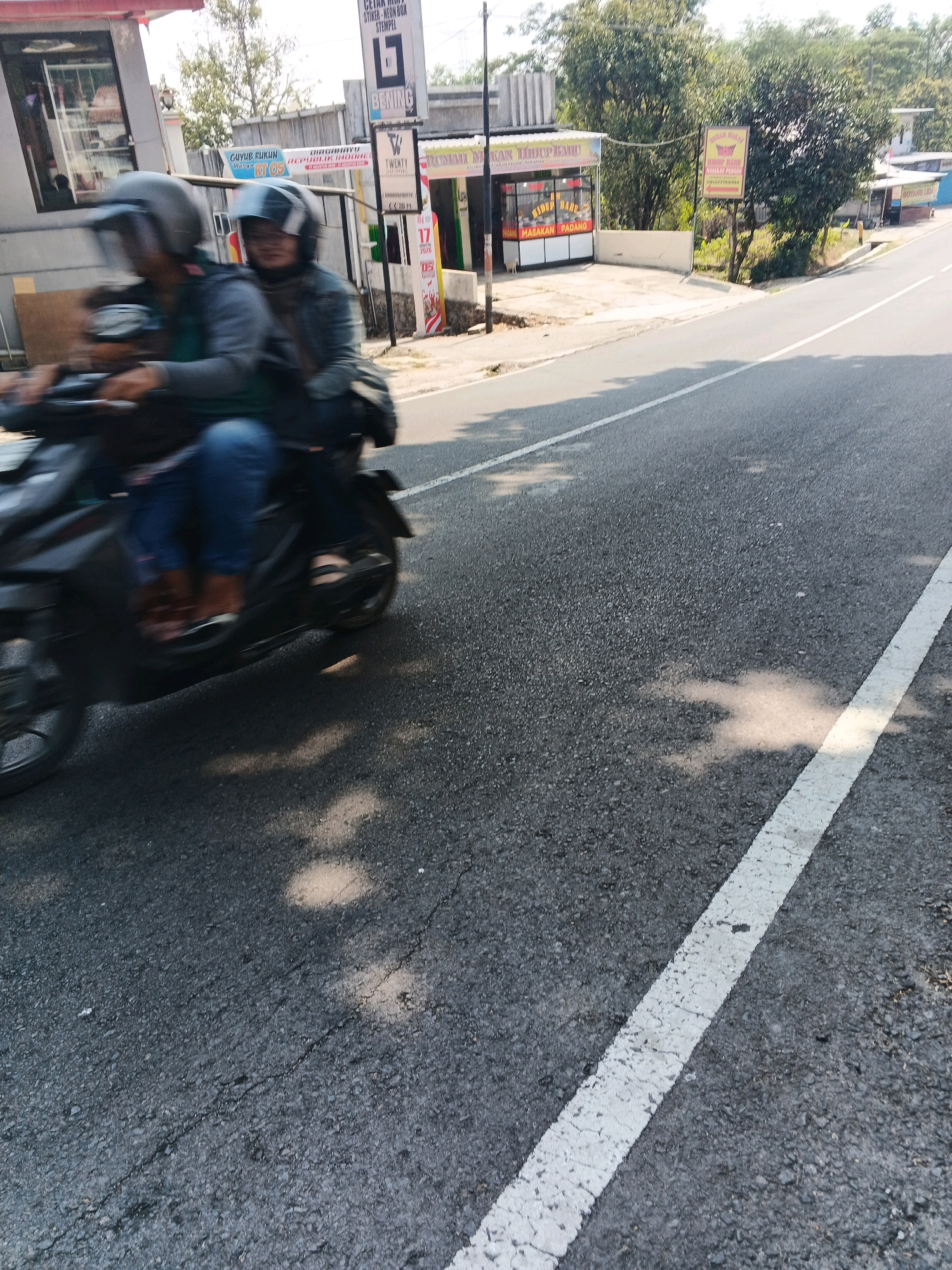 Two individuals are riding on a black motorcycle on a paved road. The driver is wearing a dark helmet and a gray jacket, while the passenger is wearing a helmet with a clear visor and a dark jacket. Both are blurred due to motion. The motorcycle is moving from left to right. The road has a white lane marking on the right side. In the background, there are buildings, trees, and signs, suggesting an urban or semi-urban environment. One sign, visible on a building, appears to advertise "Masakan Padang," a type of Indonesian cuisine, and another sign is for "Gabag Rukun." The general scene depicts everyday life in Pakopen, Indonesia, with people commuting on motorcycles.