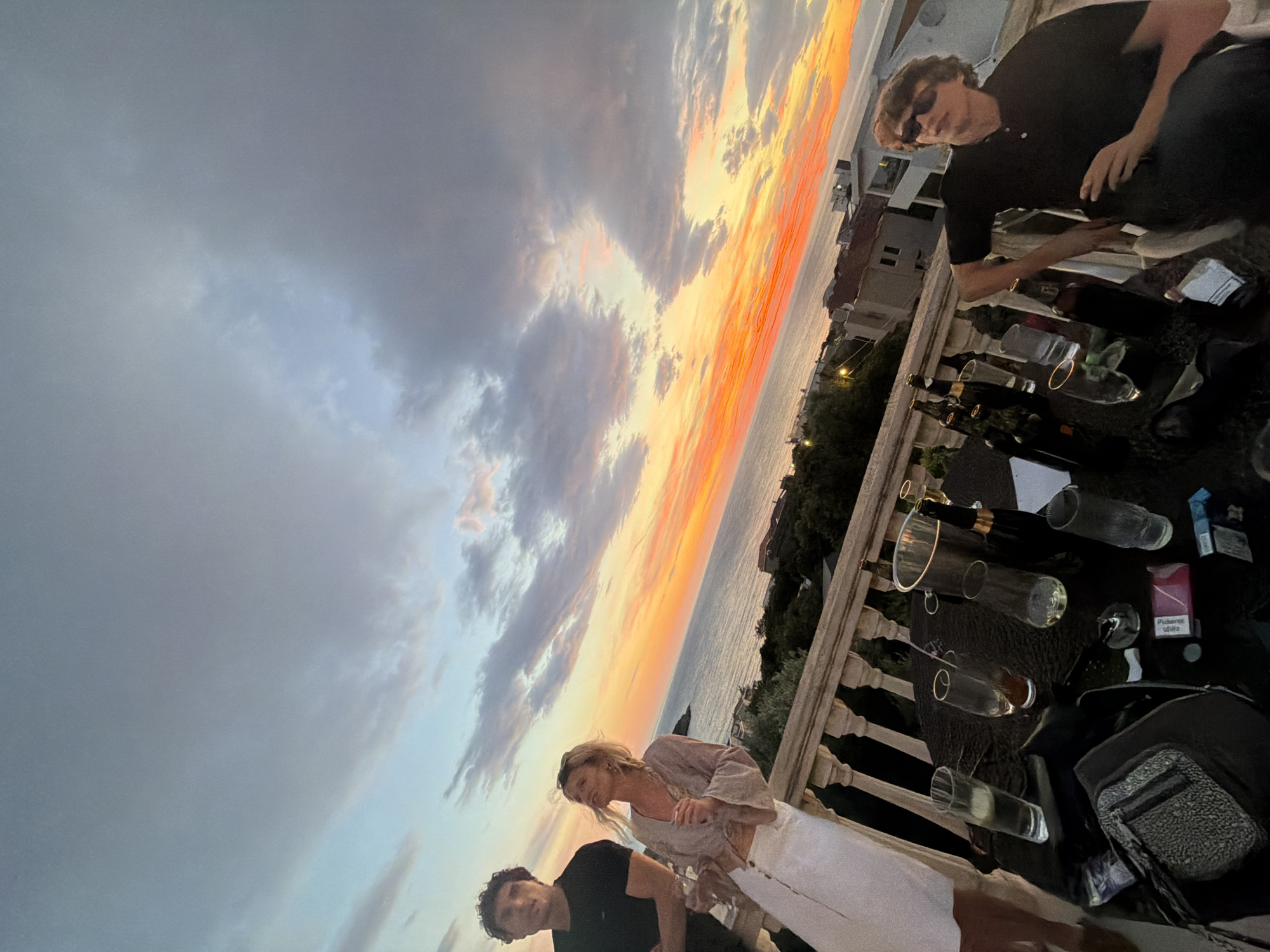The image shows a sunset in Kruče - Kërruçi, Montenegro. Three people are visible on a balcony. One man wears sunglasses and a black shirt. A woman stands with a man in a black shirt; she wears a light-colored blouse and white pants. A table is covered with empty wine bottles, glasses, cigarettes, and a handbag. The sky shows a mix of grey clouds and orange from the setting sun above the sea. Buildings are visible beyond the balcony.