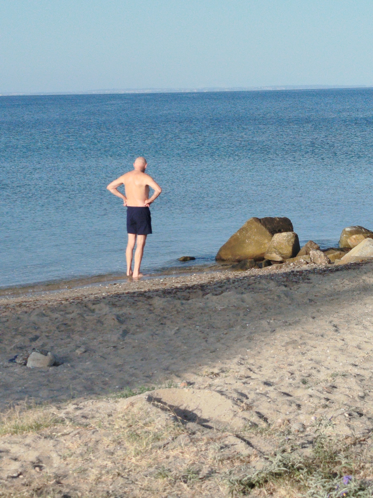 An adult male, seen from behind, stands on a sandy beach in the Sithonia Municipal Unit, Greece. He is wearing dark blue swim trunks and has his hands on his hips. The man is positioned near the water's edge, with his bare feet submerged. The sea is a calm, deep blue. In the background, large rocks are visible partially submerged in the water. The beach itself is composed of sand and small pebbles. The sky is a clear, light blue.