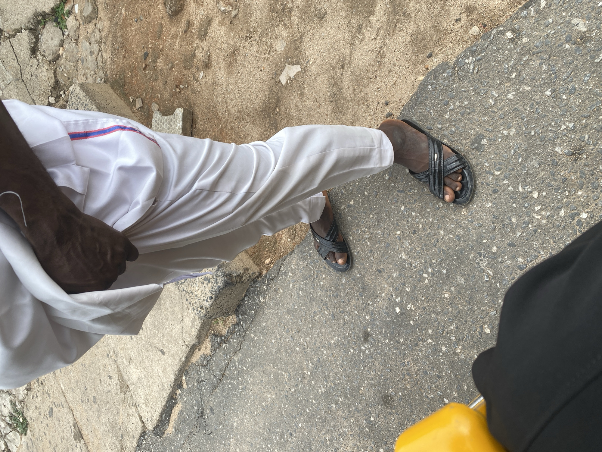This is a medium shot, taken from a high angle, showing the lower body of a person wearing a white, flowing garment and black sandals. The person is standing on a paved surface that appears to be a road or sidewalk. The ground is a mix of asphalt and concrete, with some debris scattered around. The lighting suggests it is daytime, possibly overcast, as there are no strong shadows. The overall scene is mundane, possibly a street in Jalingo, Nigeria, where the person might be walking or waiting. There is no discernible text in the image.