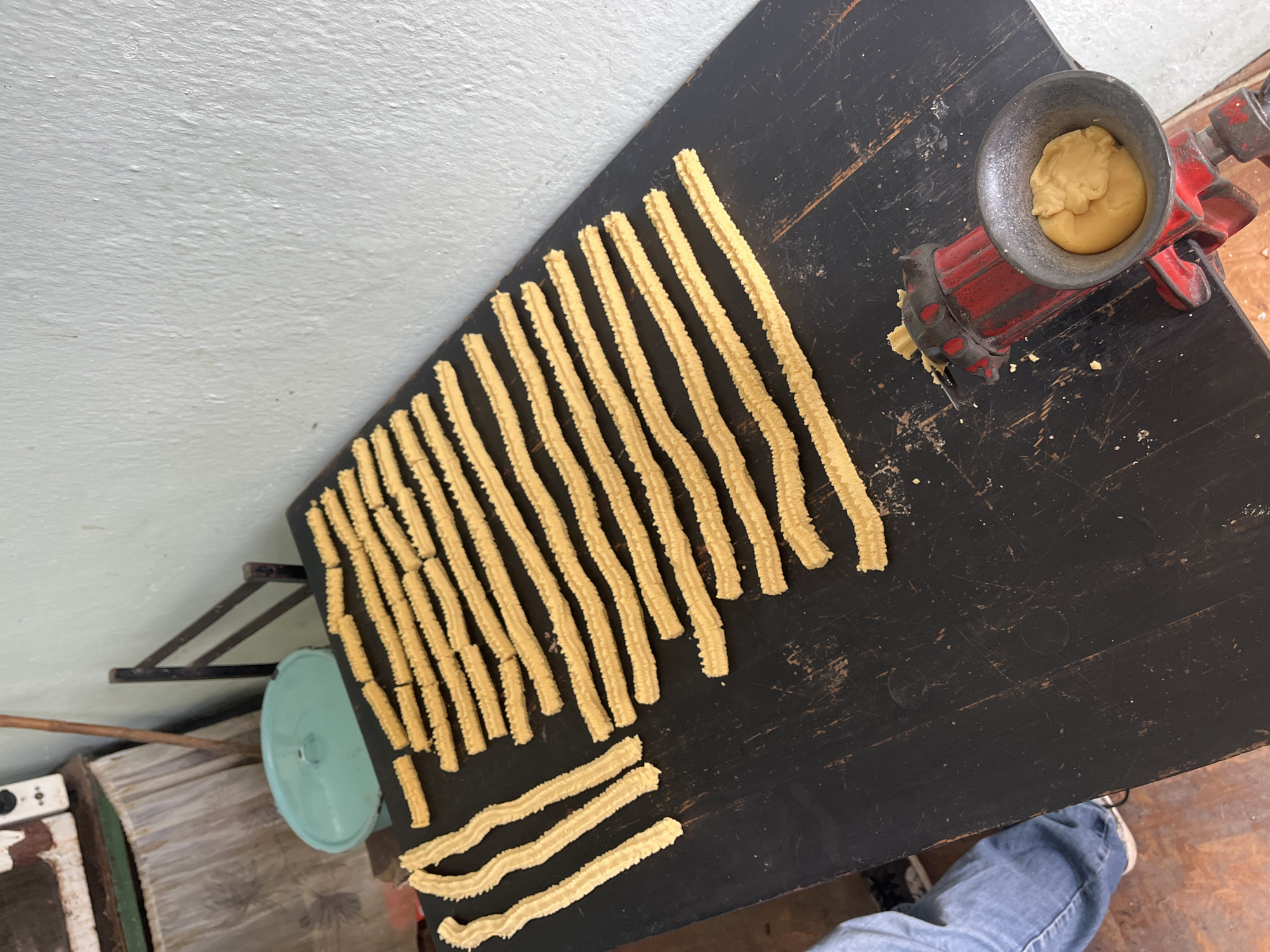 The image shows a tabletop in Hrishteni, Bulgaria, with numerous long, ridged, yellowish dough strips arranged in rows. A red, hand-cranked meat grinder-like device with a gray bowl filled with more dough is positioned on the right side of the table. A turquoise metal object, possibly a pan lid, sits on a shelf below the table, along with other miscellaneous items, including a green and white object. A section of a person's leg in blue jeans and a white sneaker is visible in the lower right corner. The wall in the background is light gray.
