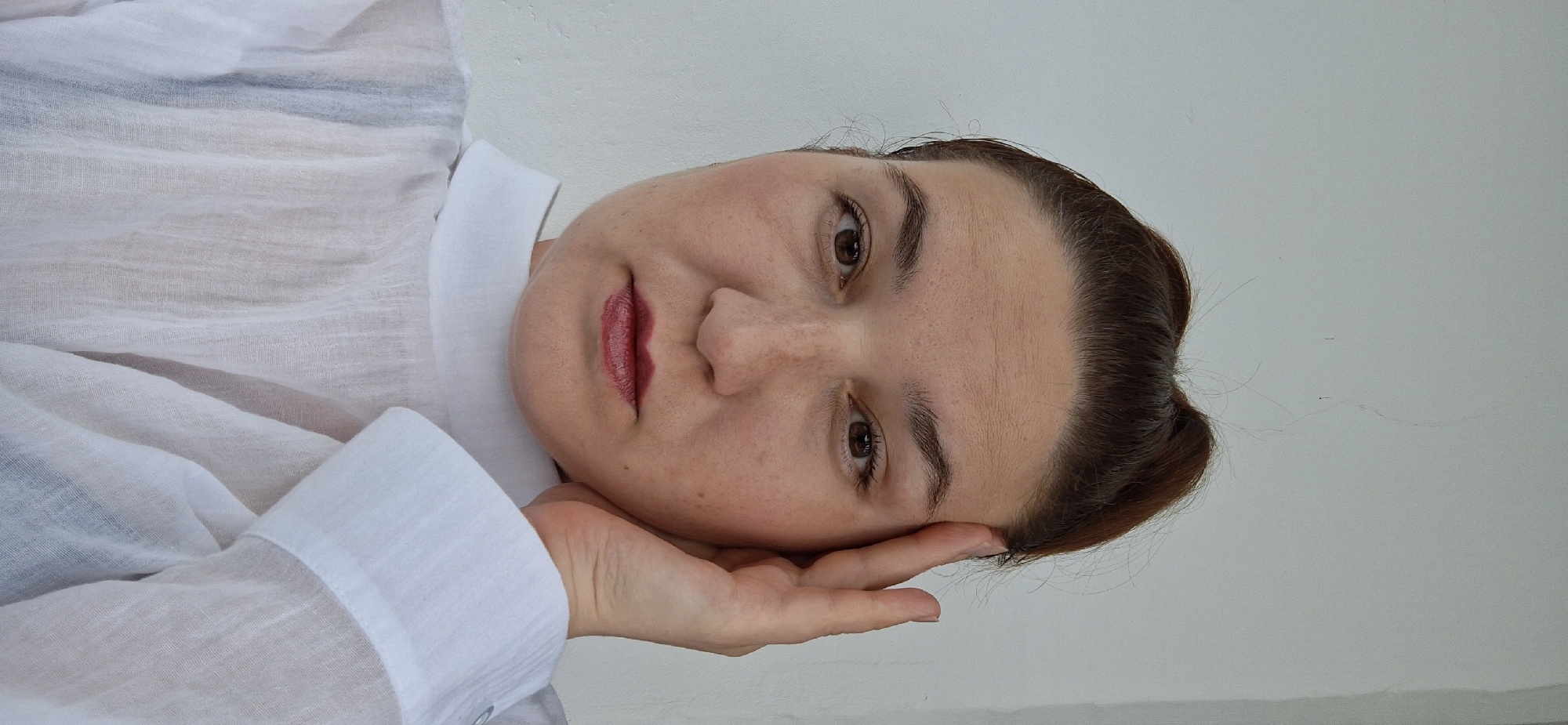 A woman with light skin, wearing a white shirt, is resting her head on her hand. Her hair is brown and pulled back. She has brown eyes and is wearing red lipstick. The background is a plain white wall. The location is the city of Chaikovsky, Russia.