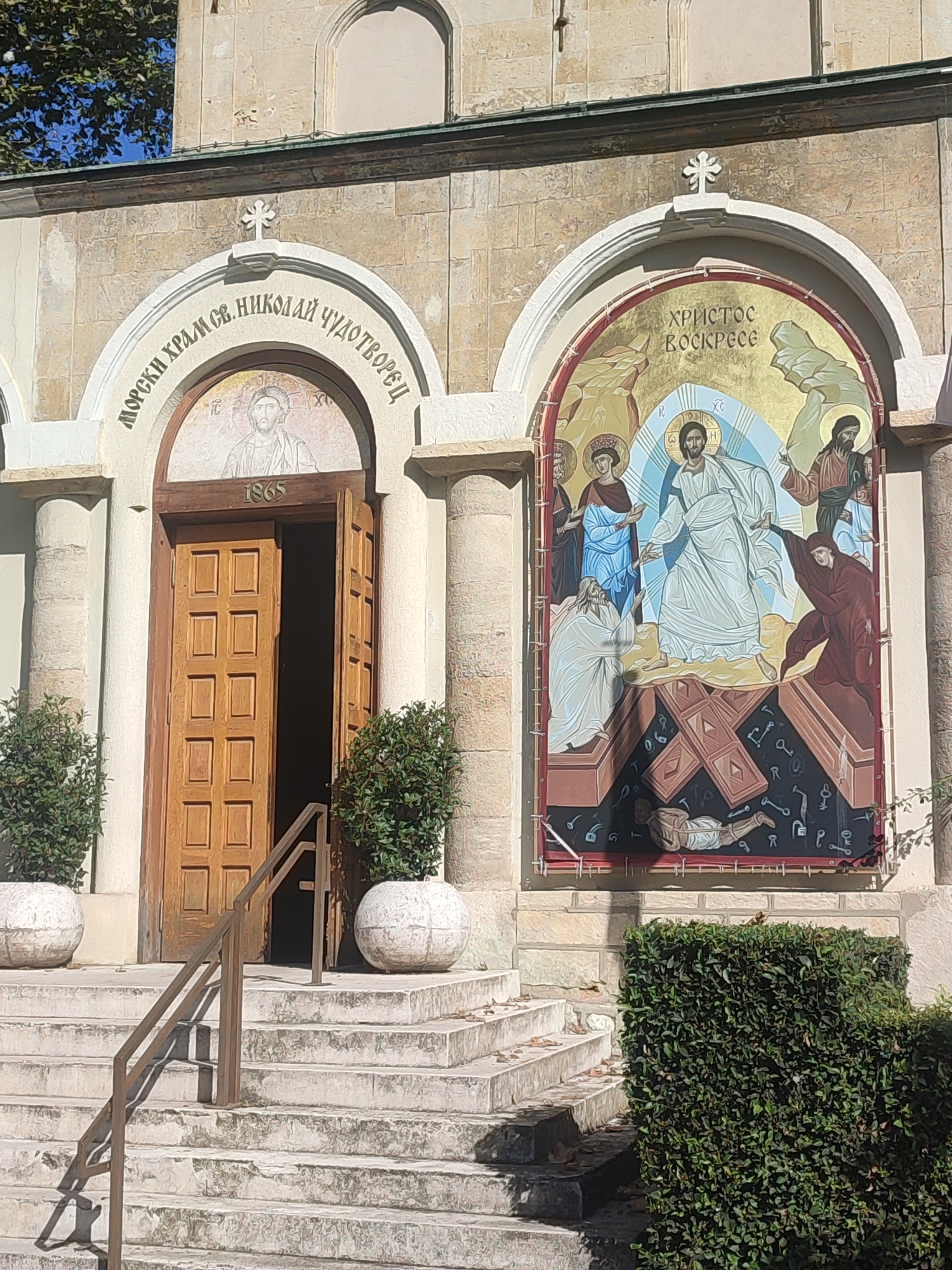 The image shows the entrance to the "Saint Nicholas the Wonderworker" Maritime Church in Varna, Bulgaria. The church facade is made of stone blocks. Above the wooden double doors, an arched mosaic depicts a portrait of Jesus Christ and the inscription "1865" below it. To the right of the entrance, a large mural illustrates Christ's resurrection. The mural features Jesus emerging from a tomb, with figures of mourners and disciples around him. The scene is set against a golden background. The entrance is accessed by a stone staircase with a metal handrail. Two large white planters with green shrubs flank the stairs, and a well-maintained green hedge is situated to the right of the entrance. The sky is visible above the church.