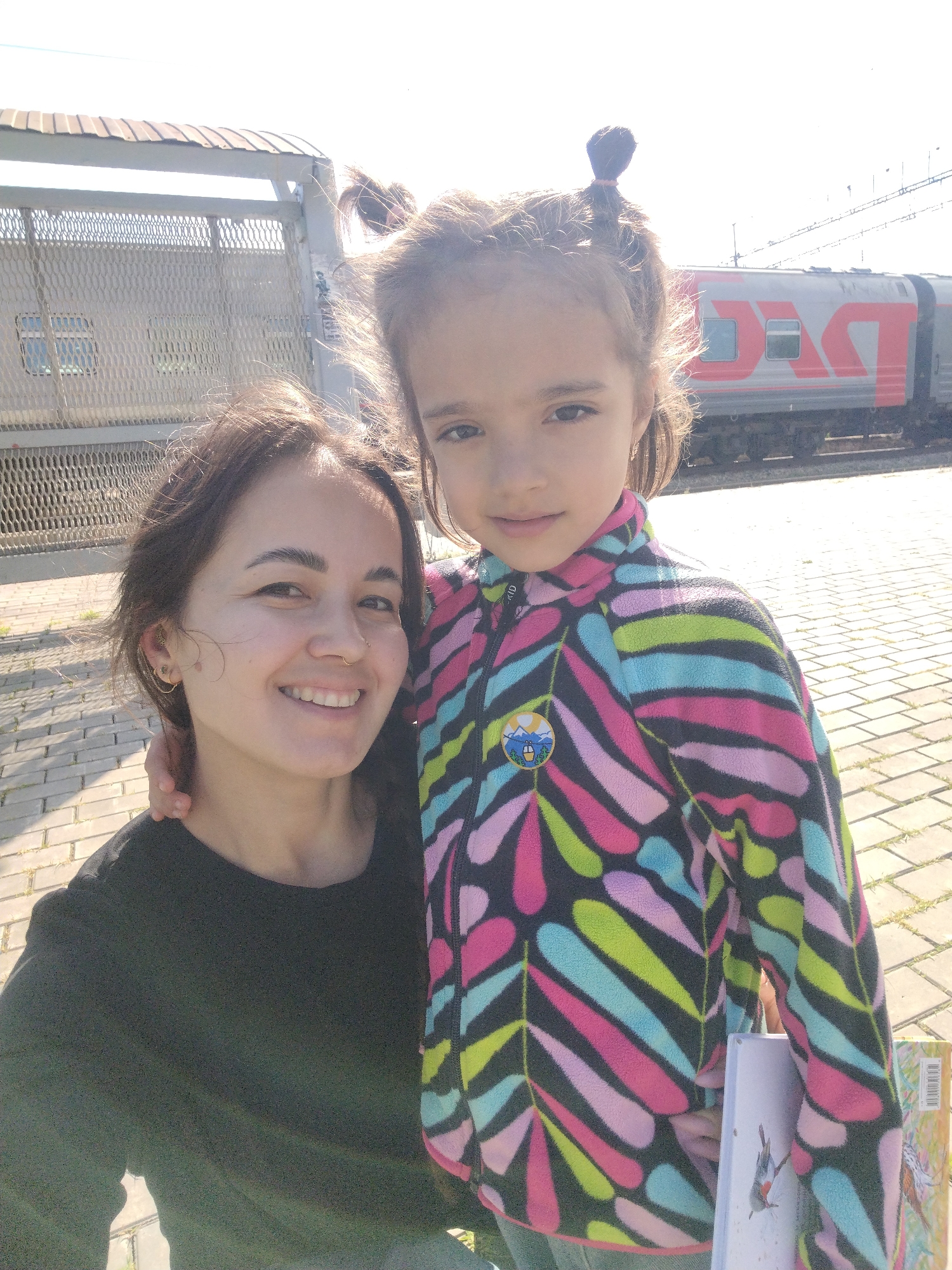 In a selfie taken outdoors, likely at a train station in Kuyeda, Russia, a woman and a young girl are positioned in the foreground. The woman, on the left, has brown hair and is smiling at the camera, wearing a black long-sleeved shirt. A small ring is visible in her nose, and she has multiple earrings in her left ear. The girl, to the right, has her hair styled in two buns and is wearing a multi-colored fleece jacket with a leaf pattern in shades of pink, blue, green, and black. She is holding a book. In the background, a train with a red logo is visible, along with a fence and a paved walkway. The sun is bright, casting shadows and creating a washed-out effect in the sky.
