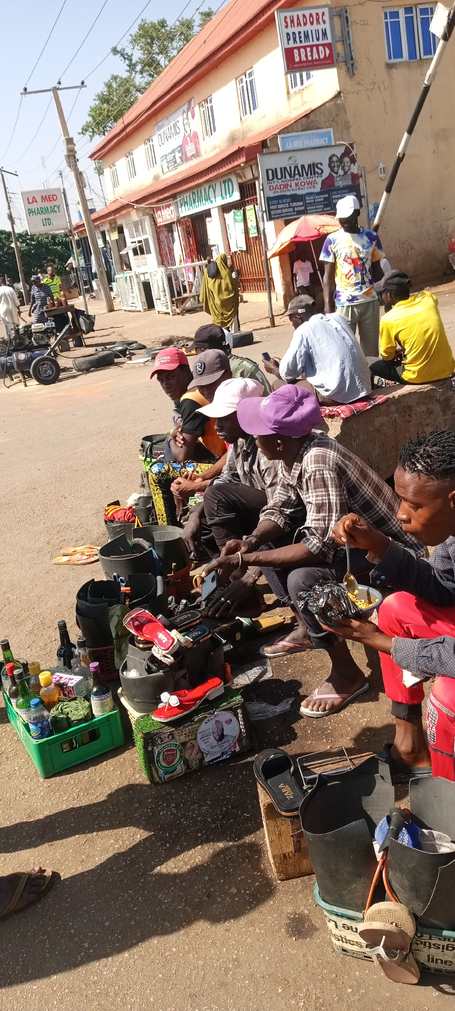 The image captures a scene in Jos, Nigeria. A row of men are seated on a low concrete wall, likely engaged in shoe repair or cleaning. Each man has his tools and materials arranged around him. One man is eating from a bowl using a spoon. A crate of bottles and shoes are visible in the lower left corner, with additional shoes and sandals near the men.
Behind the men, there is a pharmacy building with signs reading "LA MED PHARMACY LTD." and "SHADORC PREMIUM BREAD." A Dunamis International Gospel Centre sign is also visible. A few people are walking in the background. A generator is on the street in the distance.