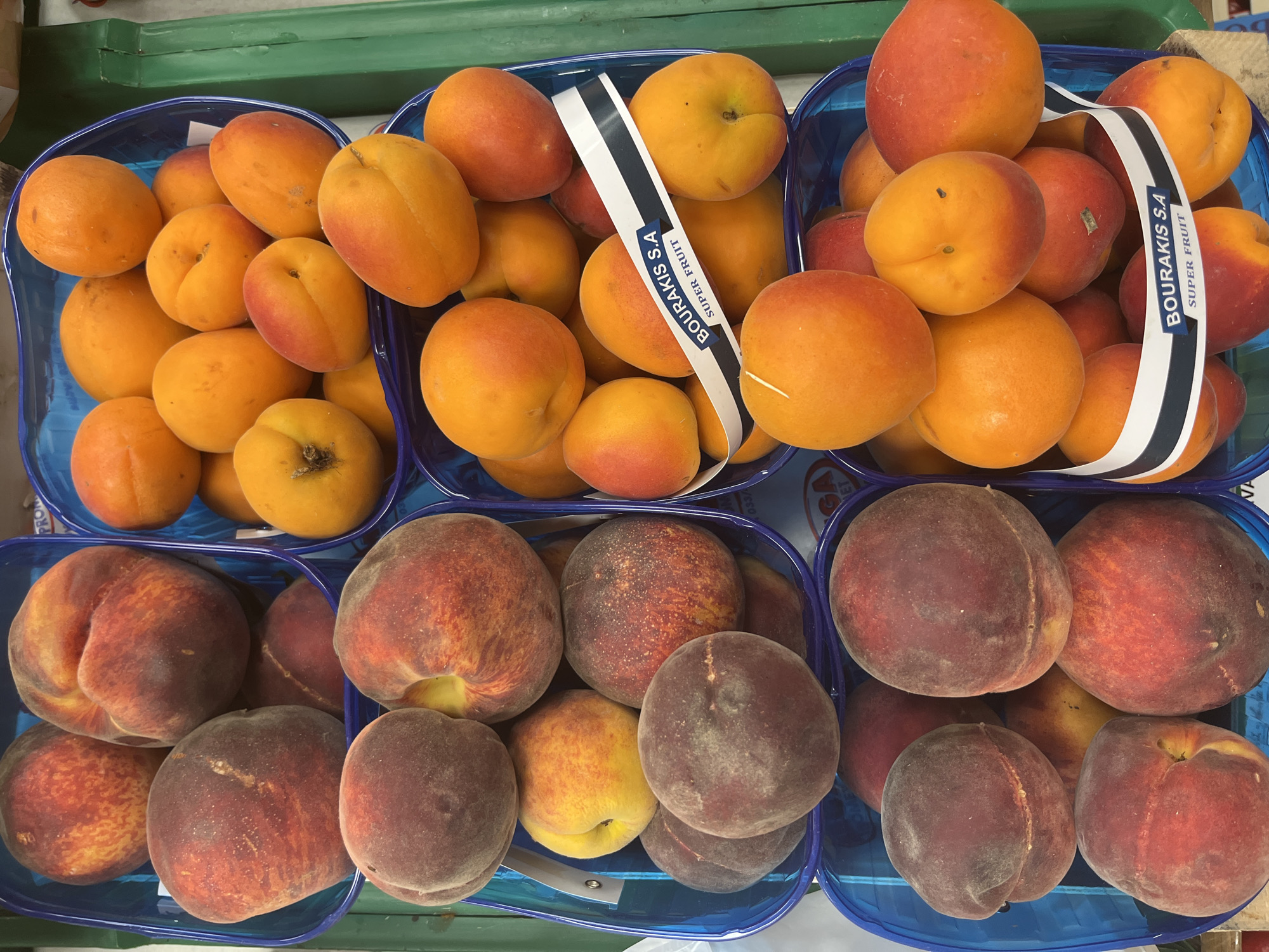 The image is a close-up, top-down view of several blue plastic crates filled with fruit. The crates are stacked and appear to be in a market or store.
The fruits in the crates are primarily peaches and apricots. The peaches are large, fuzzy, and range in color from deep red to yellow and orange. Some of the apricots are smaller and have a smooth skin, with a bright orange-red color. One of the apricot crates has a white label with blue text that reads "BOURAKIS S.A. SUPER FRUIT".
The background is slightly blurred but appears to be the interior of a retail environment with hints of green shelving or walls. The lighting is bright, suggesting artificial market lighting. There are no people or animals visible in the image. The context of Budva, Montenegro, is not discernible from the image itself.
