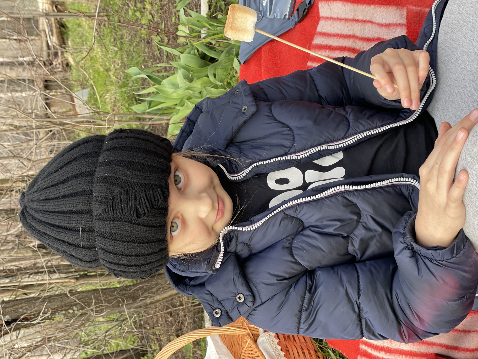 A young child wearing a black knit hat and a navy blue puffer jacket sits outdoors, holding a stick with a toasted marshmallow on it. The child has fair skin and green eyes and is smiling slightly at the camera. The background shows a red and white striped blanket, foliage, and trees, indicating an outdoor setting. A basket is partially visible in the lower left corner. There is no information to confirm the location as Балдейка, Russia.