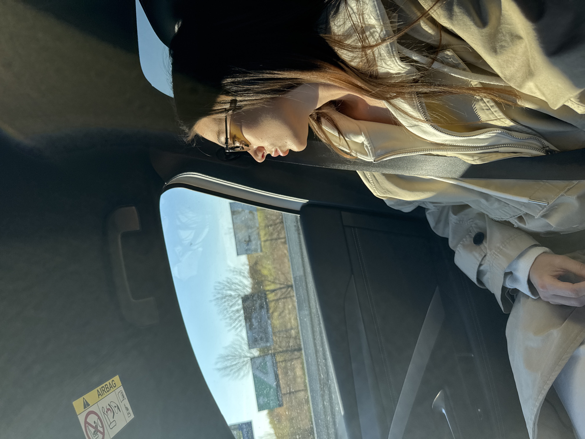 The image shows a person wearing glasses and a tan jacket in a car. The person's face is in profile, and they are looking out the window. The car is moving, and the view outside the window includes a light blue sky and trees that have lost their leaves in the winter. The image seems to be taken at a time that there may be a light dusting of snow or ice on the ground or on the trees. The location is Nieporęt, Poland.