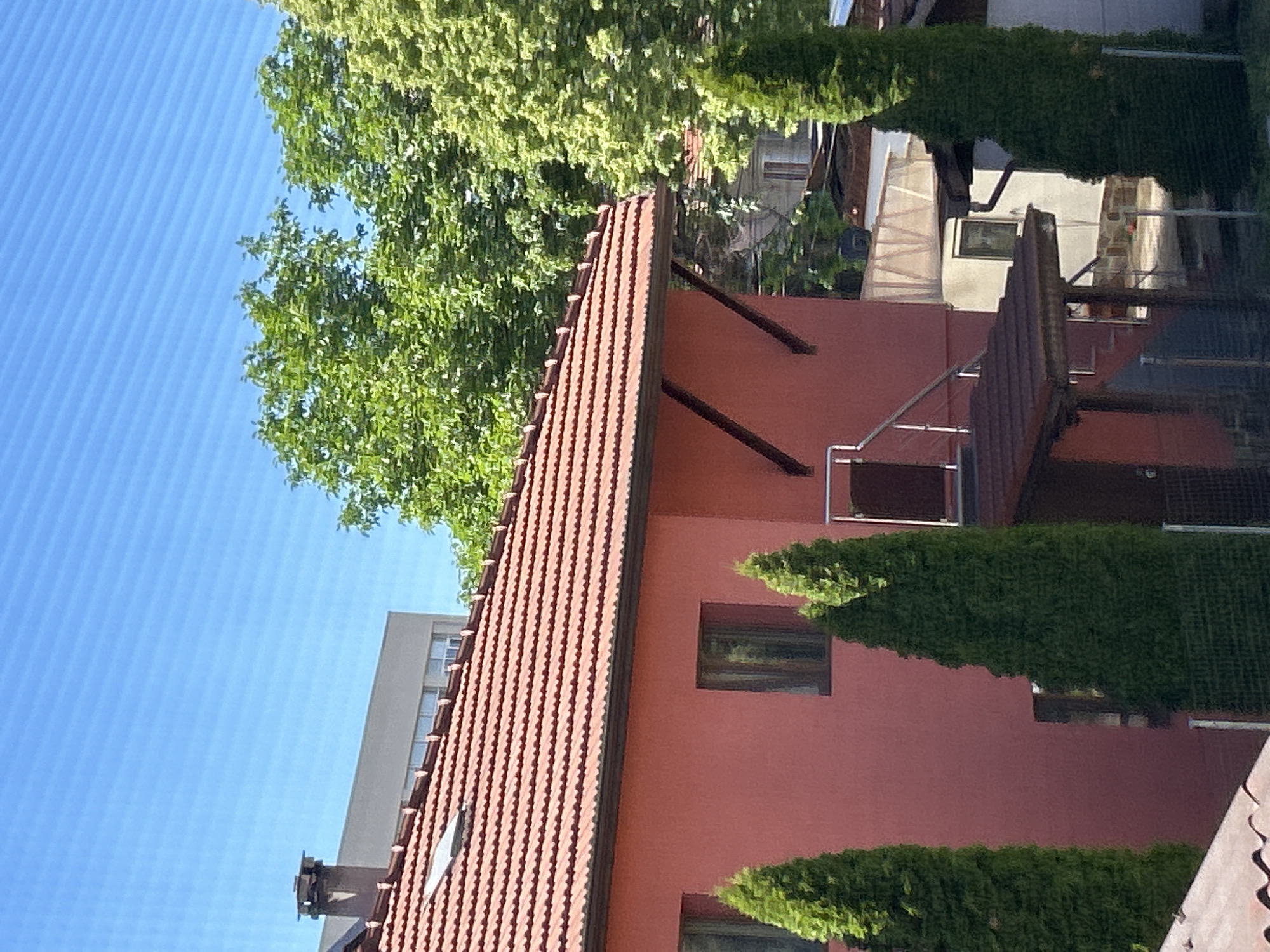 The image shows a residential building in Sofia, Bulgaria, characterized by a red tile roof and reddish-brown facade. The sky is blue and visible to the left of the building, partially obscured by green foliage, possibly trees. Adjacent to the building is a light gray structure, likely another building. Conical-shaped shrubs are present near the building's facade. A staircase with metal railings is visible on the building's side.