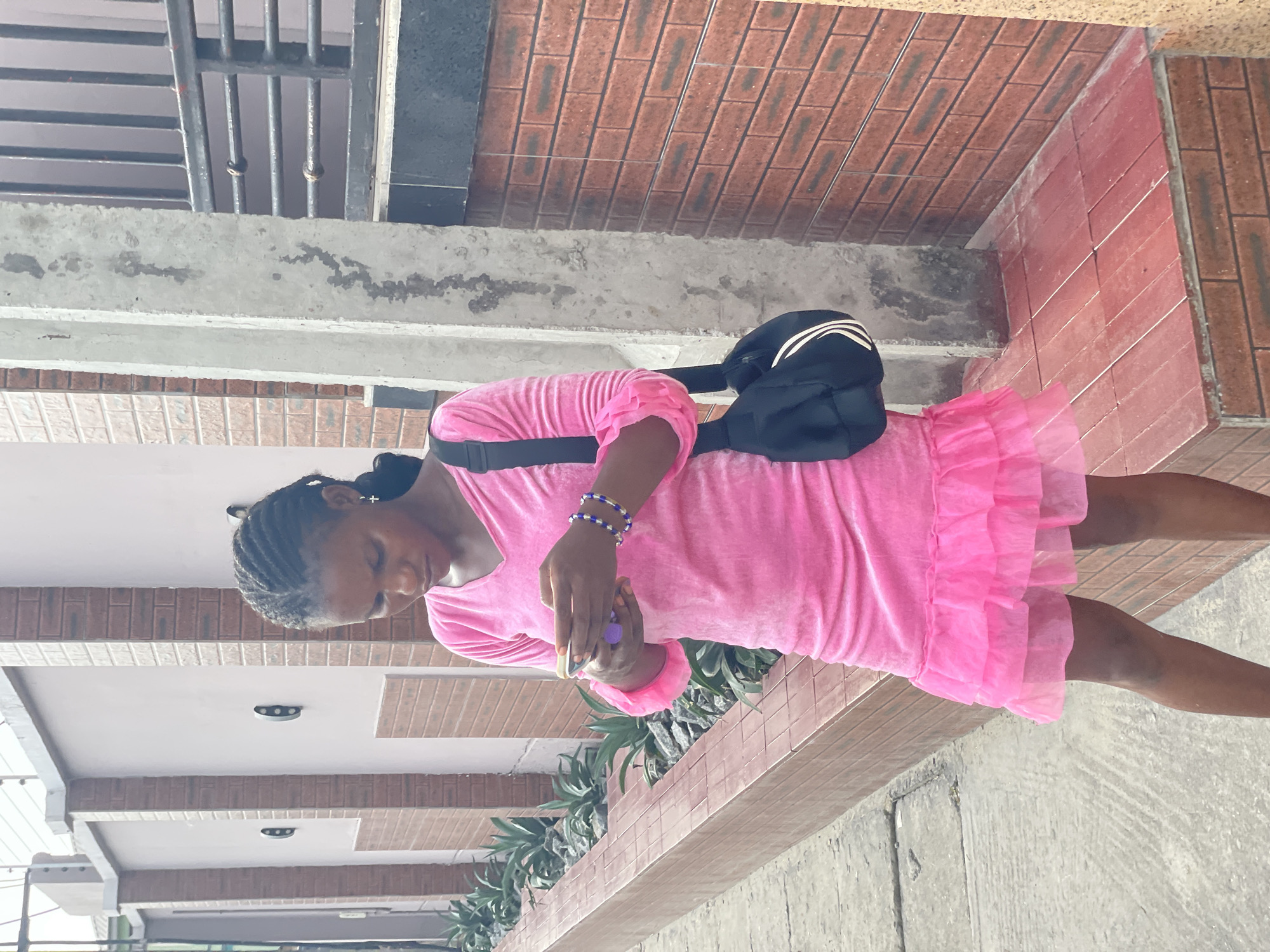 A young woman stands outdoors next to a building in Iranla, Nigeria, captured in a brightly lit daytime setting. She is positioned facing slightly left, her gaze directed downwards at a small purple object held in her hands, suggesting focus or engagement. She has dark braided hair and wears a vibrant pink dress with a velvet-like upper part and a ruffled, sheer skirt. Several blue and white beaded bracelets adorn her right wrist, and a black fanny pack with white stripes is slung across her body.
The background reveals a building facade characterized by reddish-brown brickwork and grey concrete structures, some showing signs of weathering. Along a concrete ledge, there are planters filled with green, leafy plants. Higher up, a metal security grate or window bars are visible. The ground beneath her is a concrete surface. The overall lighting is strong and even, indicating a sunny day. No visible text is present in the image.
