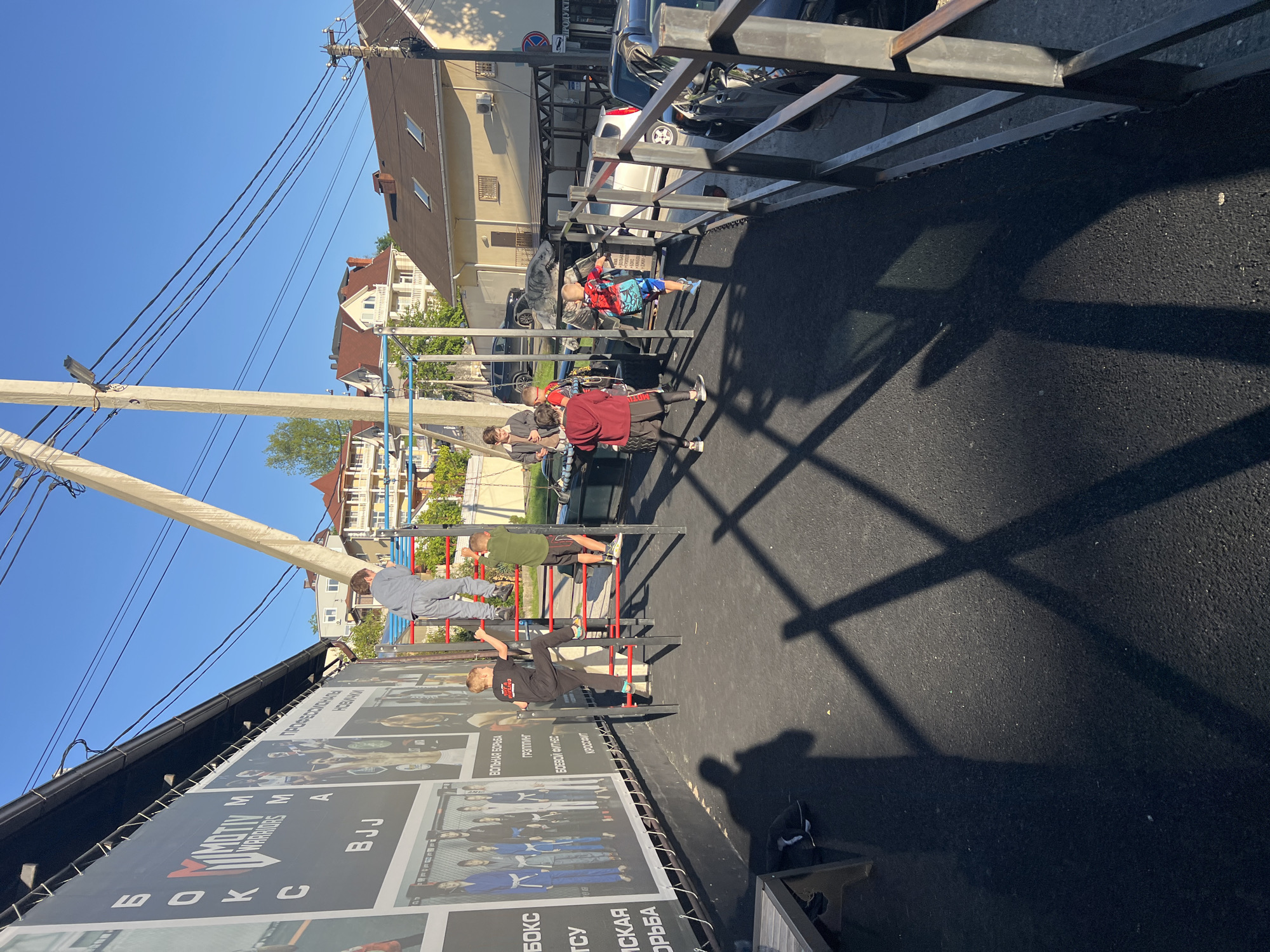 The image captures an outdoor calisthenics or street workout area in Sochi, Russia, under bright, clear conditions. The scene is dominated by several individuals, predominantly young males or children, actively engaged with various metal exercise structures.
In the foreground, a large billboard stretches along the side of a building, displaying imagery related to martial arts and fitness. Visible text on the billboard includes "MIMOTIV WARRIORS," "BJJ," "БОКС" (Boxing), "ВОЛЬНАЯ БОРЬБА" (Freestyle Wrestling), "КРОССФИТ" (CrossFit), "РУЗИТИН," and "ПРОФЕССИОНАЛЬНАЯ" (Professional) and "НОВЫЙ" (New).
On the asphalt ground, distinct shadows from the exercise equipment and overhead utility lines are cast by the strong sunlight, indicating it is likely midday or early afternoon with a clear blue sky. A tall utility pole with numerous wires runs across the upper left portion of the image.
The main subjects are the people using the equipment. One boy, dressed in a red and blue top with a Spiderman-like design and blue shorts, lies face down on a lower metal frame. Nearby, another person in a dark red hoodie and black pants stands beside a large tire. Further along the exercise setup, a person in a grey tracksuit is climbing a red and black ladder-like structure. Another individual in a green t-shirt and dark shorts is also ascending a similar structure. A person in black athletic wear is shown in the process of climbing another section of the multi-level metal bars.
In the background, behind the exercise area, parked cars are visible, along with multi-story buildings featuring tiled roofs, suggesting a residential or mixed-use urban environment. A partially obscured road sign in the distance includes the word "ПРОДУКТЫ" (Groceries). The overall impression is of an active community space dedicated to physical training.