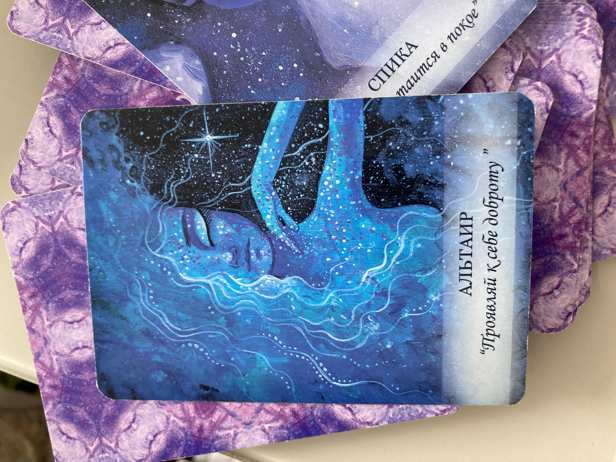 The image shows a stack of tarot-like cards. The top card prominently displays an artistic rendering of a person submerged in water or a watery substance. The artwork features shades of blue and white, with white dots resembling stars or sparkles scattered across the image. The person's face is visible, eyes closed, and the artwork conveys a sense of serenity or dreaminess. To the right of the image, the word "АЛЬТАИР" (ALTAIR) is written, followed by a phrase in Cyrillic script enclosed in quotation marks, "Проявляй К себе доброту" (Proyavlyay K sebe dobrotu), which translates to "Show kindness to yourself." A card behind the main card has the word "СПИКА" (SPIKA). The rest of the cards have a purple patterned design. The location in the city of Florianópolis, Brazil is unknown.
