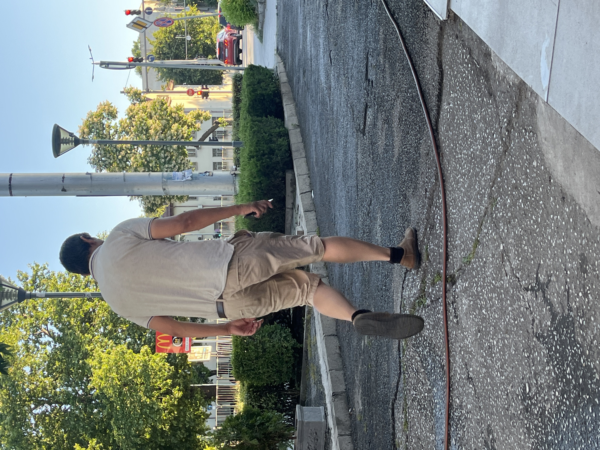 The image depicts a man walking away from the camera on a cracked asphalt path in Plovdiv, Bulgaria. He is wearing a light-colored polo shirt, khaki shorts, dark socks, and brown shoes. The man is holding what appears to be a cigarette in his right hand. In the background, there is a McDonald's restaurant sign, a tall light pole, a traffic light, and buildings. Green trees and hedges are visible, suggesting an urban setting during daytime. A red hose lies across the path. The scene captures a mundane moment in a city environment.