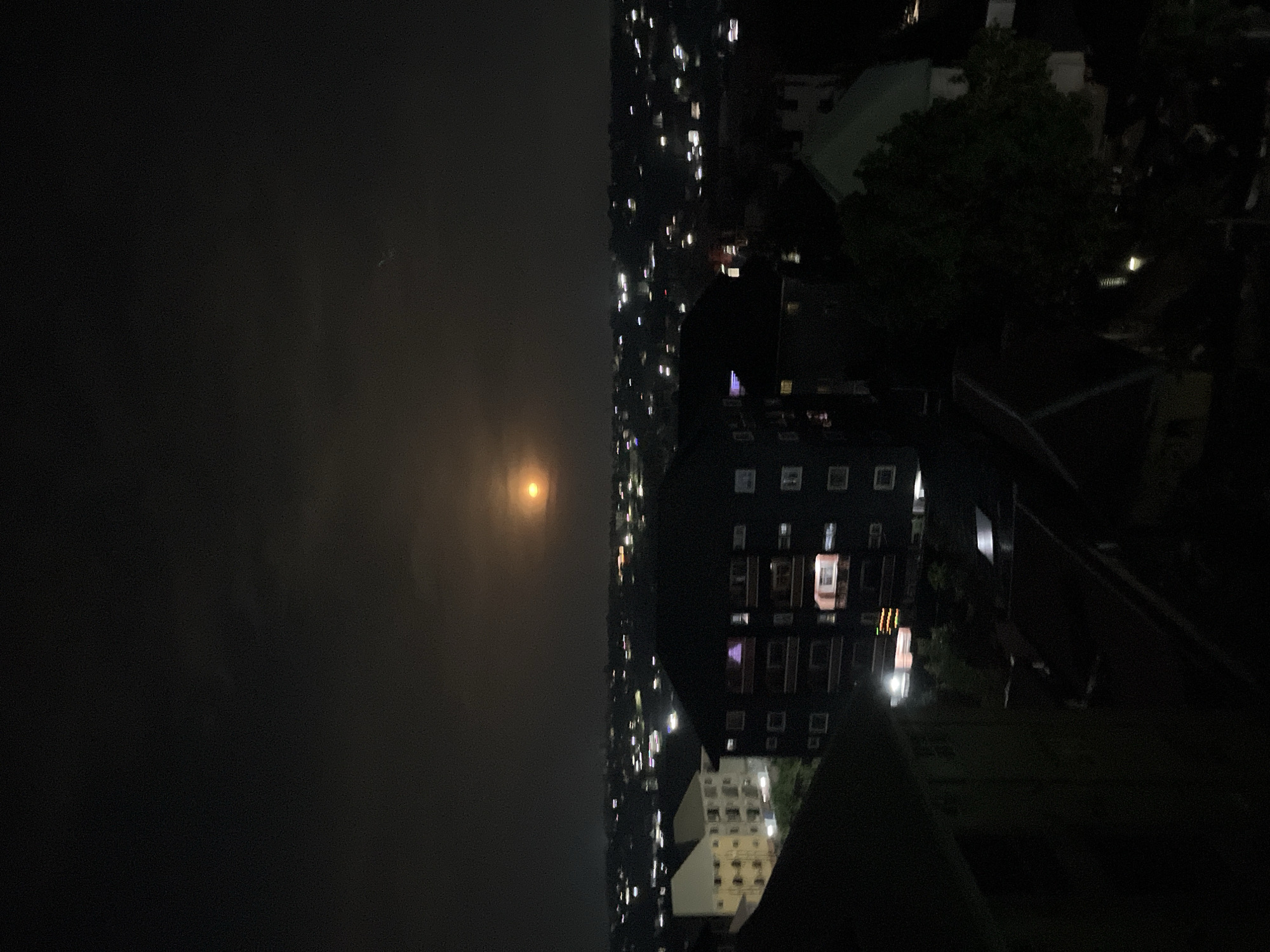 A nighttime view from an elevated position shows a section of Nkpor, Nigeria, shrouded in a hazy, low-visibility atmosphere. The skyline is punctuated by numerous lights suggesting a densely populated urban area. A light-emitting disc is seen in the sky. To the right, the image is dominated by buildings, with multiple illuminated windows suggesting residential or commercial activity. Dark forms possibly represent trees and surrounding structures, further adding to the urban texture. The overall scene exhibits a sense of nighttime urbanity.