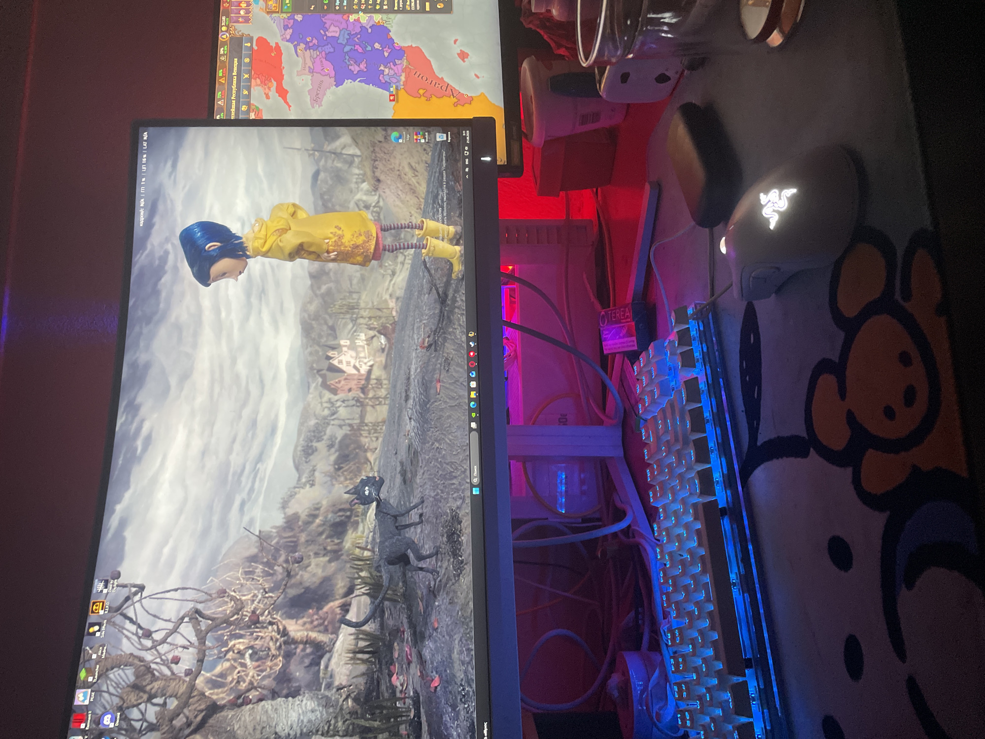 The image shows a computer setup with two monitors. The left monitor displays a scene from the movie "Coraline," featuring the character Coraline in her yellow raincoat and a black cat. The right monitor shows a map-based game interface with colorful regions and text in Russian. In the foreground, a white keyboard with illuminated blue keys and a white gaming mouse with a glowing green logo are visible on a desk mat. Cables are neatly managed behind the monitor. The environment appears to be a home or office space, with reddish lighting in the background. The provided location details of Neoi Epivates, Greece, cannot be visually confirmed from the image.