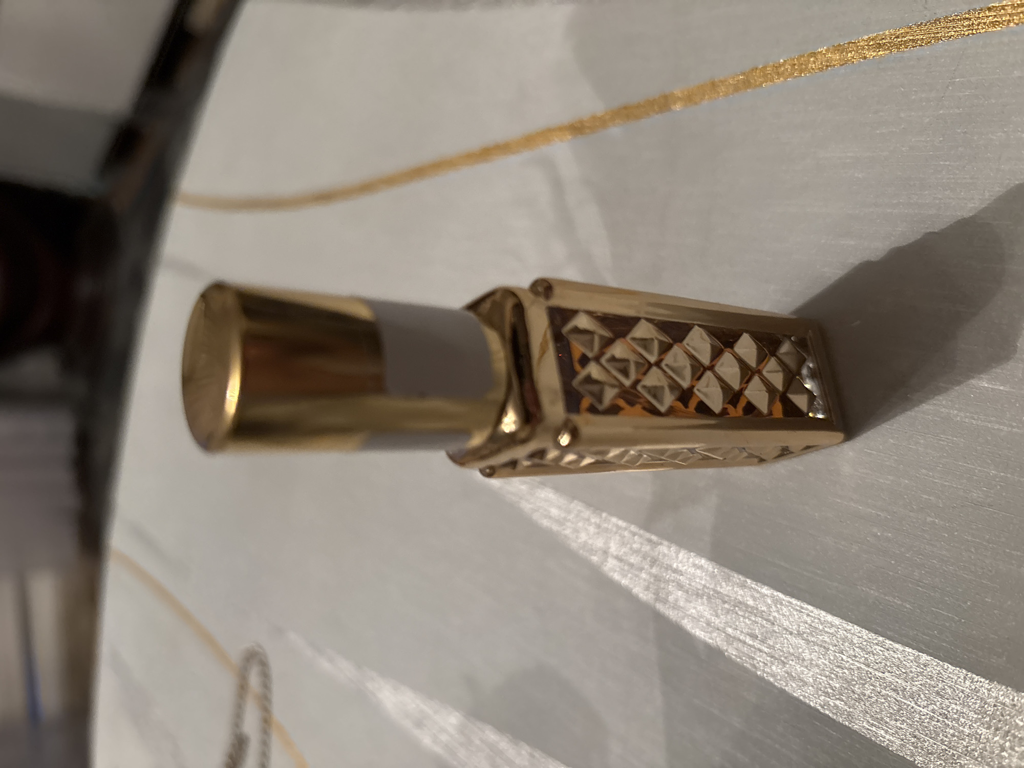 The image shows a close-up, angled view of a small, ornate golden perfume bottle. The bottle has a cylindrical, metallic gold cap on one end, which transitions into a rectangular body. The body of the bottle is decorated with a repeating diamond pattern in a darker, rich tone, possibly amber or brown, inset within the gold frame. A thin, textured gold ribbon or stripe runs diagonally across the background of the image, with another similar stripe of shimmering silver material below it, creating a textured and reflective surface. The lighting suggests it might be daytime, with strong shadows cast by the bottle, indicating a light source from the side. The setting appears to be indoors, on a decorative surface. There is no visible text in the image. The overall impression is one of luxury and elegance due to the gold and diamond-like embellishments.