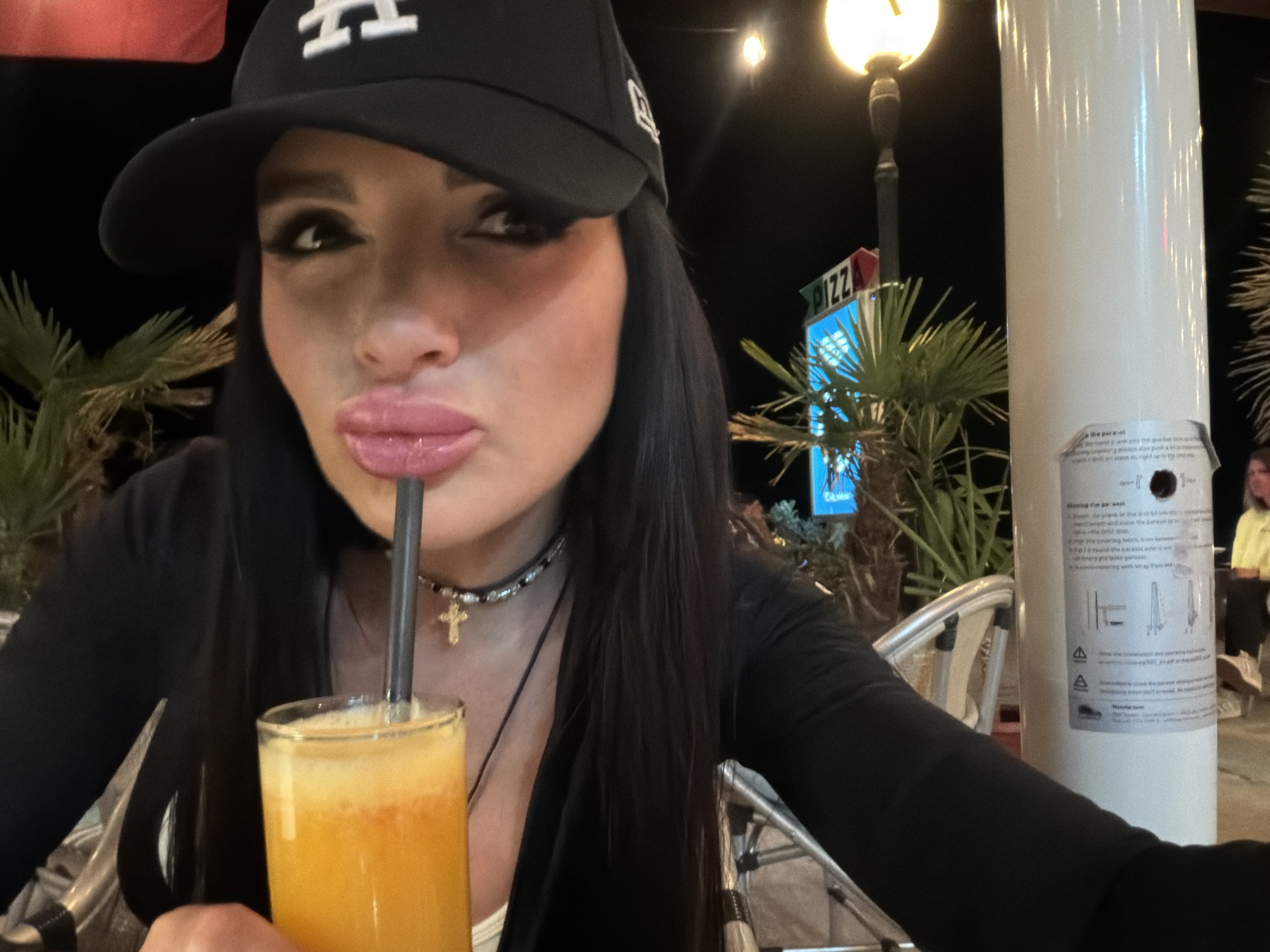 A close-up selfie captures a woman holding a glass of orange beverage with a black straw to her full lips, seemingly taking a drink at night. She has long dark hair, and is wearing a black baseball cap with white stylized letters (resembling "LA"), a black long-sleeved top over a white inner top, and a beaded choker necklace with a small gold cross pendant. Her face is made up, with noticeable eyeliner/eyeshadow.
The scene is an outdoor cafe or restaurant patio at night in Rafailovići, Montenegro. The background is dark, illuminated by an ornate streetlamp on the left and artificial lighting on signs. Green, palm-like plants are visible. Metal-framed outdoor seating can be partially seen. To the right, a prominent white cylindrical pole or column features a white sticker with black text and diagrams, providing instructions for "the parasol," with words like "open," "close," "installation," and "manufacturer" discernable. Behind the pole, another person in a yellow top is partially visible. A blue sign in the mid-background clearly displays "PIZZA" in red and white letters, with other indistinct text below it. The atmosphere appears casual and relaxed.