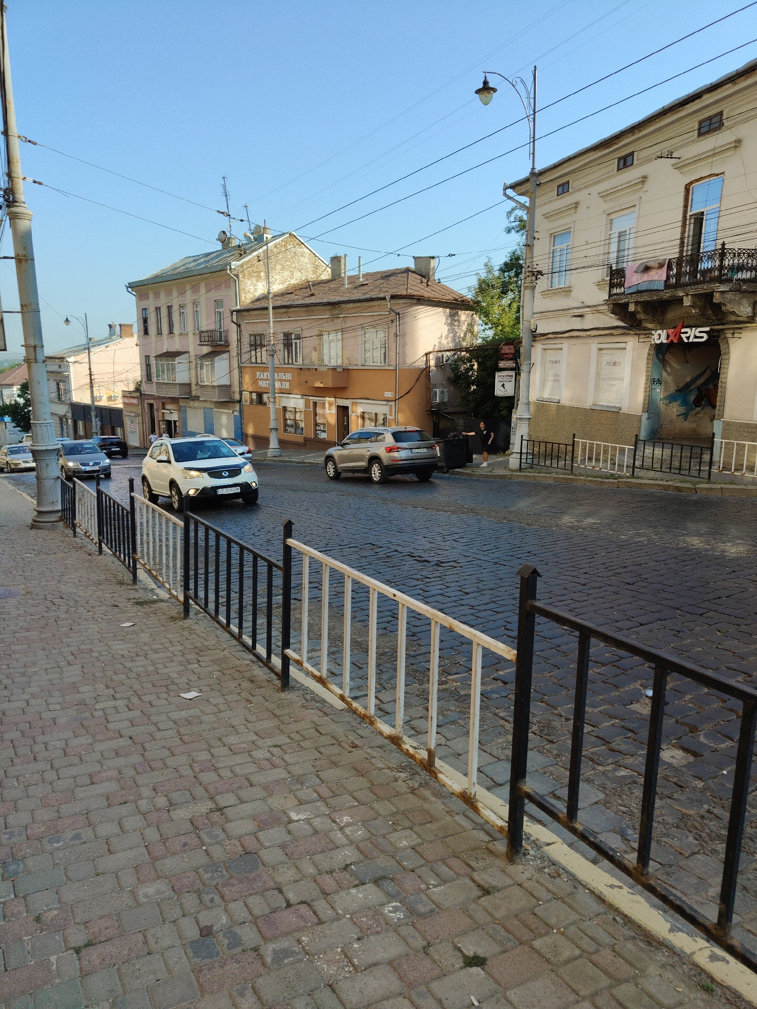 The image shows a street scene in Chernivtsi, Ukraine, likely on a sunny day given the clear blue sky. The street is paved with cobblestones, and there are buildings lining both sides. On the right side of the street, a large, multi-story building with a cream-colored facade is visible. This building appears to have shops or businesses on the ground floor, with signs that are partially legible, including "POLARIS". There's a decorative balcony on the upper floor.
On the left side of the street, a sidewalk is visible, also paved with cobblestones. A metal fence separates the sidewalk from the street, featuring a combination of black and white painted metal posts and railings. There are several cars driving on the street, including a white SUV in the foreground, and another car partially visible further down the street. A pedestrian, dressed in dark clothing, is standing near a large dark trash receptacle on the right side of the street. The overall impression is of a historic urban setting with active street life.