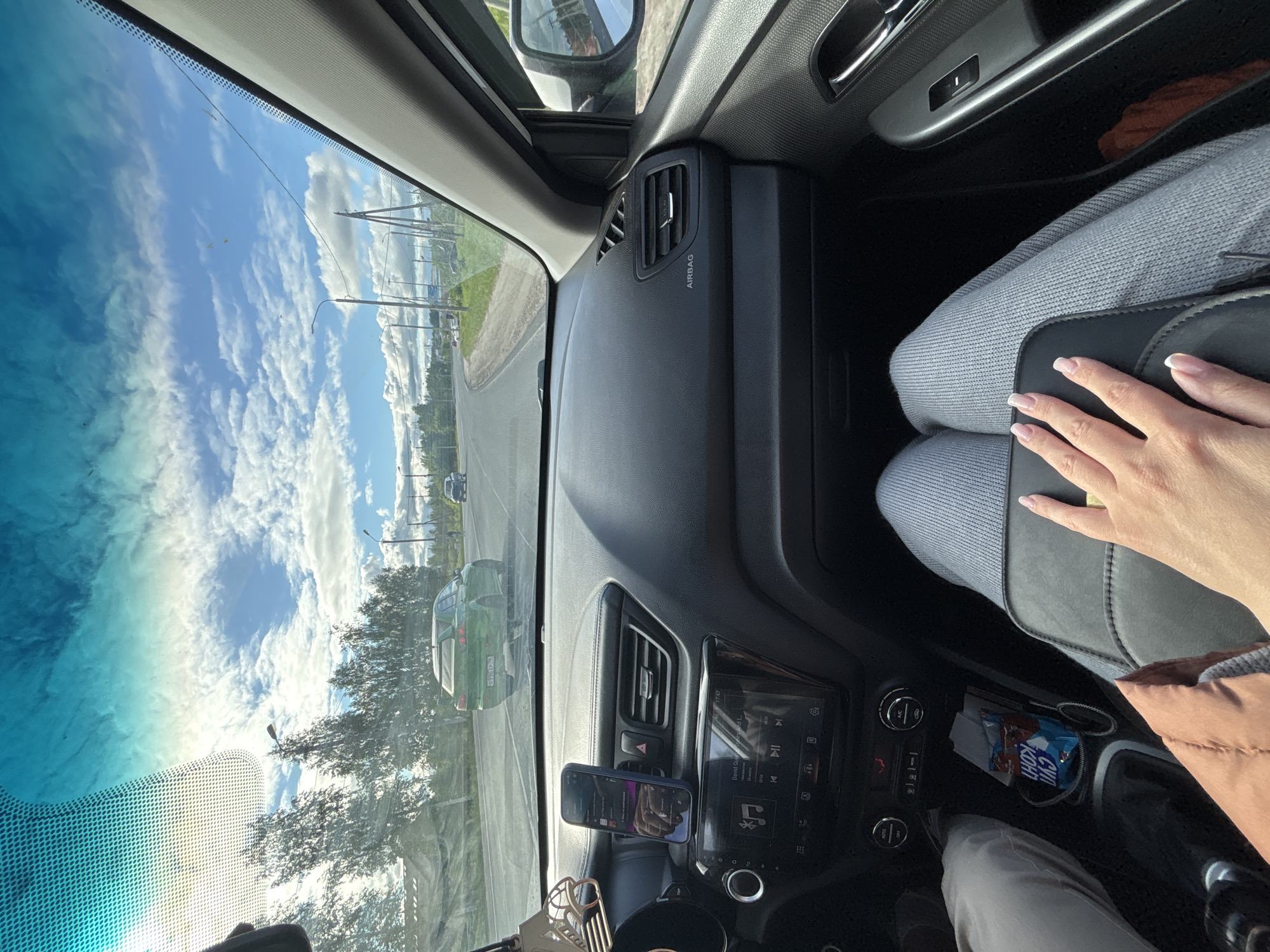 A view from inside a car driving on a sunny day. The sky is visible through the windshield, showing white clouds and blue sky. The road is visible, with a green car in the adjacent lane. The dashboard of the car is visible, with a mobile phone mounted in a holder, and the car's infotainment system displaying music information. The passenger's legs are visible, with their hand resting on the seat. A chocolate bar wrapper and an aux cable are in a compartment in the center console. The location appears to be Yaroslavl, Russia, based on the architecture visible through the windshield.