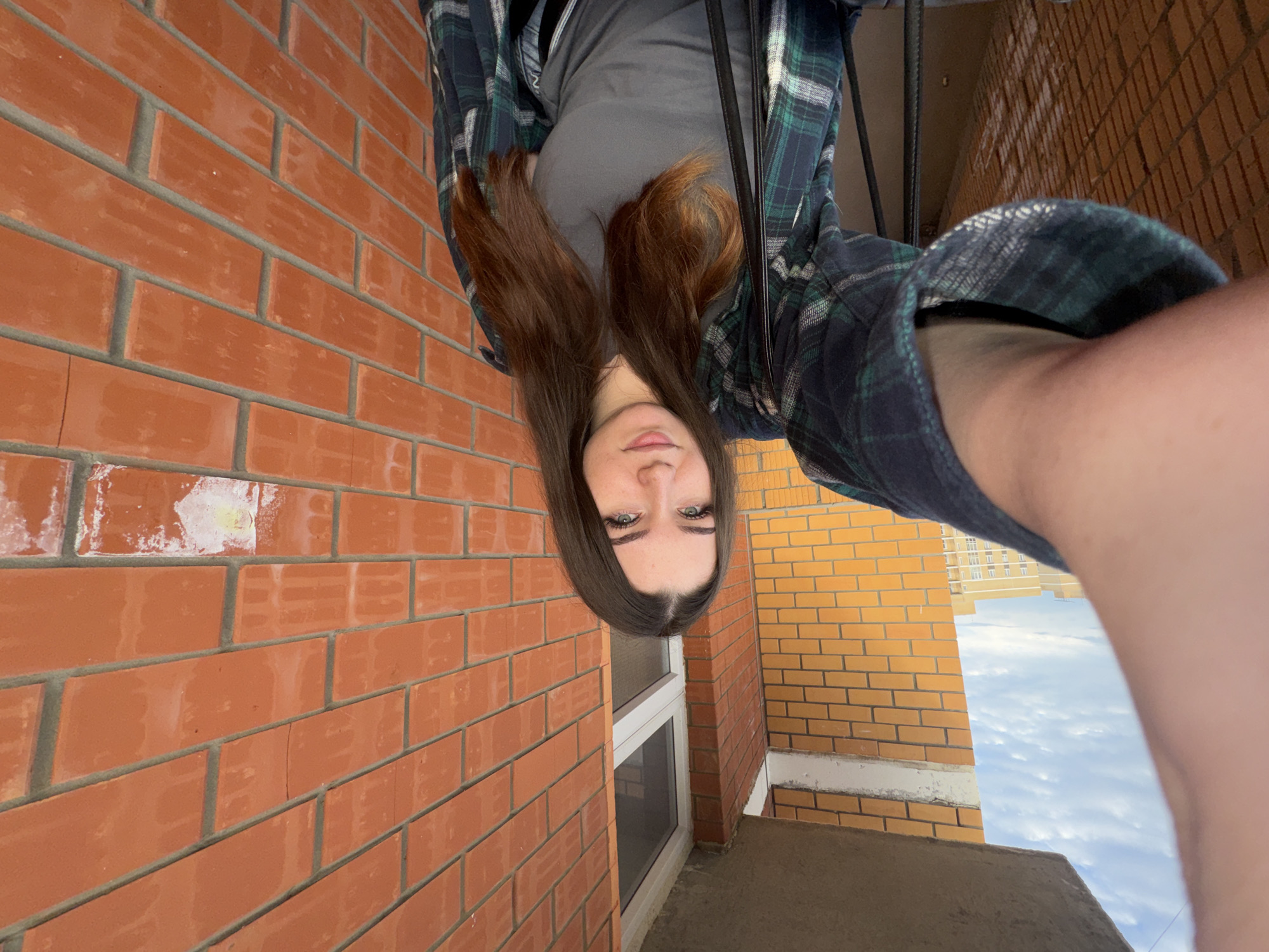 The media file shows an upside-down, close-up selfie of a young woman. She has long, dark brown hair, green eyes, and fair skin. She is wearing a grey t-shirt and an open, dark green and blue plaid shirt over it. The background is a brick wall, suggesting an outdoor setting. To the right of the woman, there's a glimpse of a building with yellow brick and a cloudy sky. The image appears to have been taken during the daytime, given the natural light. The woman's expression is neutral. No text is visible in the image. The context provided indicates the location is Perm, Russia.