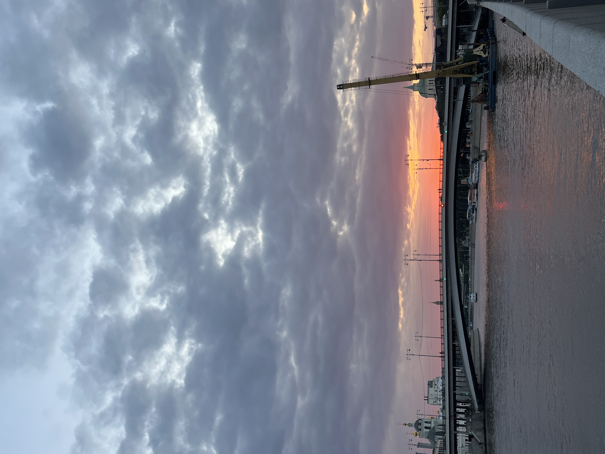 The image depicts a view of a bridge over a river at sunset in Moscow, Russia. The sky is filled with dramatic clouds, with the sun setting on the horizon, casting a warm, orange and pink glow. The bridge, a large concrete structure, spans the width of the river and is illuminated by streetlights. Several boats are visible on the water, some docked and others moving. A large construction crane is positioned on a barge in the foreground, suggesting ongoing work or repairs near the bridge. In the background, the city skyline is visible, with architectural landmarks including domed buildings and other structures. The overall scene conveys a sense of urban activity against the backdrop of a vibrant sunset.