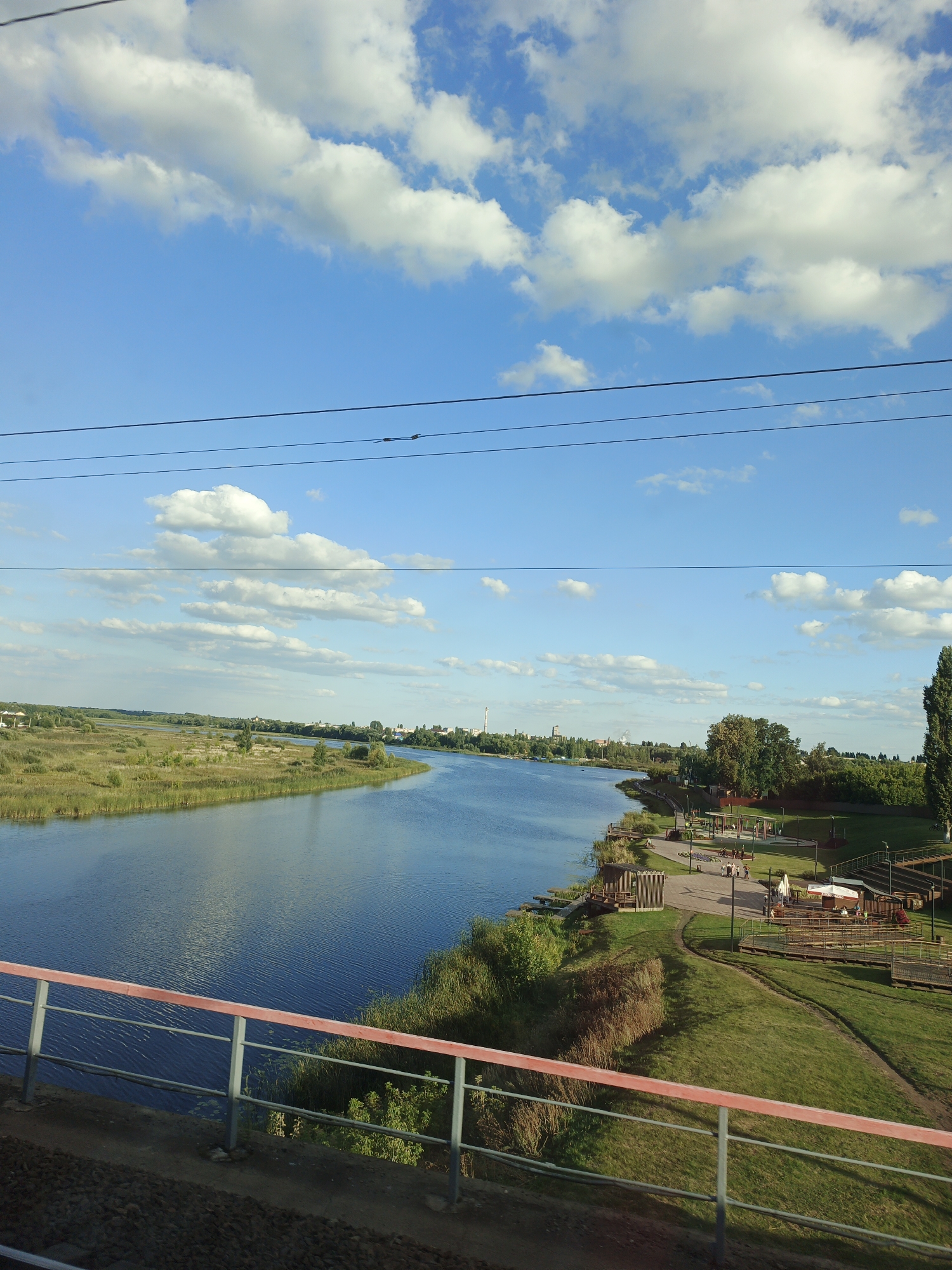 The image depicts a serene riverside scene. A wide, calm river dominates the left side of the frame, reflecting the blue sky and scattered white clouds. The riverbanks are lined with lush green grass and vegetation, with reeds growing at the water's edge on the left.

On the right bank, a well-maintained park area is visible. There are paved pathways, grassy slopes, and several wooden structures, including benches, small pavilions, and an amphitheater-like seating area. People are scattered throughout the park, some walking, others sitting and conversing. Trees of varying sizes, including tall, slender ones and broader, leafy ones, dot the landscape. In the distance, across the river and along the far bank, a town or city skyline is visible, with buildings and industrial structures, including a tall chimney, silhouetted against the sky.

The foreground is partially obscured by a red-topped railing, suggesting the image was taken from a bridge or elevated walkway. Overhead, power lines are visible, strung across the sky. The overall impression is one of a peaceful day in a developed area with natural beauty. This scene is likely located in Gryazi, Russia, given the presence of a river, park, and urban elements consistent with the city's geography.