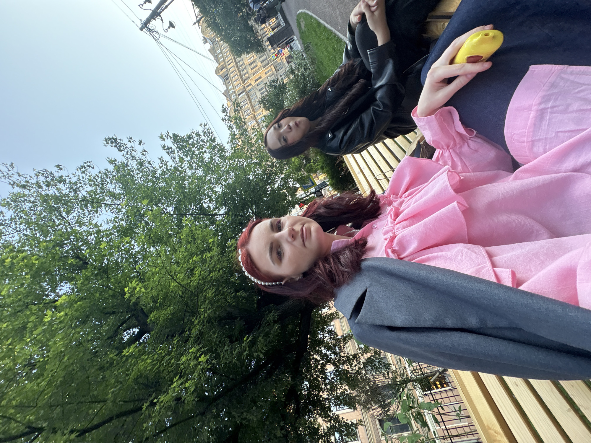 Two young women are sitting on a wooden bench in a park in Saint Petersburg, Russia. The woman on the left has red hair and is wearing a pink ruffled blouse and a gray jacket. The woman on the right has long dark hair and is wearing a black leather jacket and black pants. She is holding a yellow object that appears to be a toy. Behind them, there is a large tree, a sidewalk, and a street with buildings in the background. The sky is clear and blue. The buildings are old and have intricate architectural details. It appears to be a sunny day.