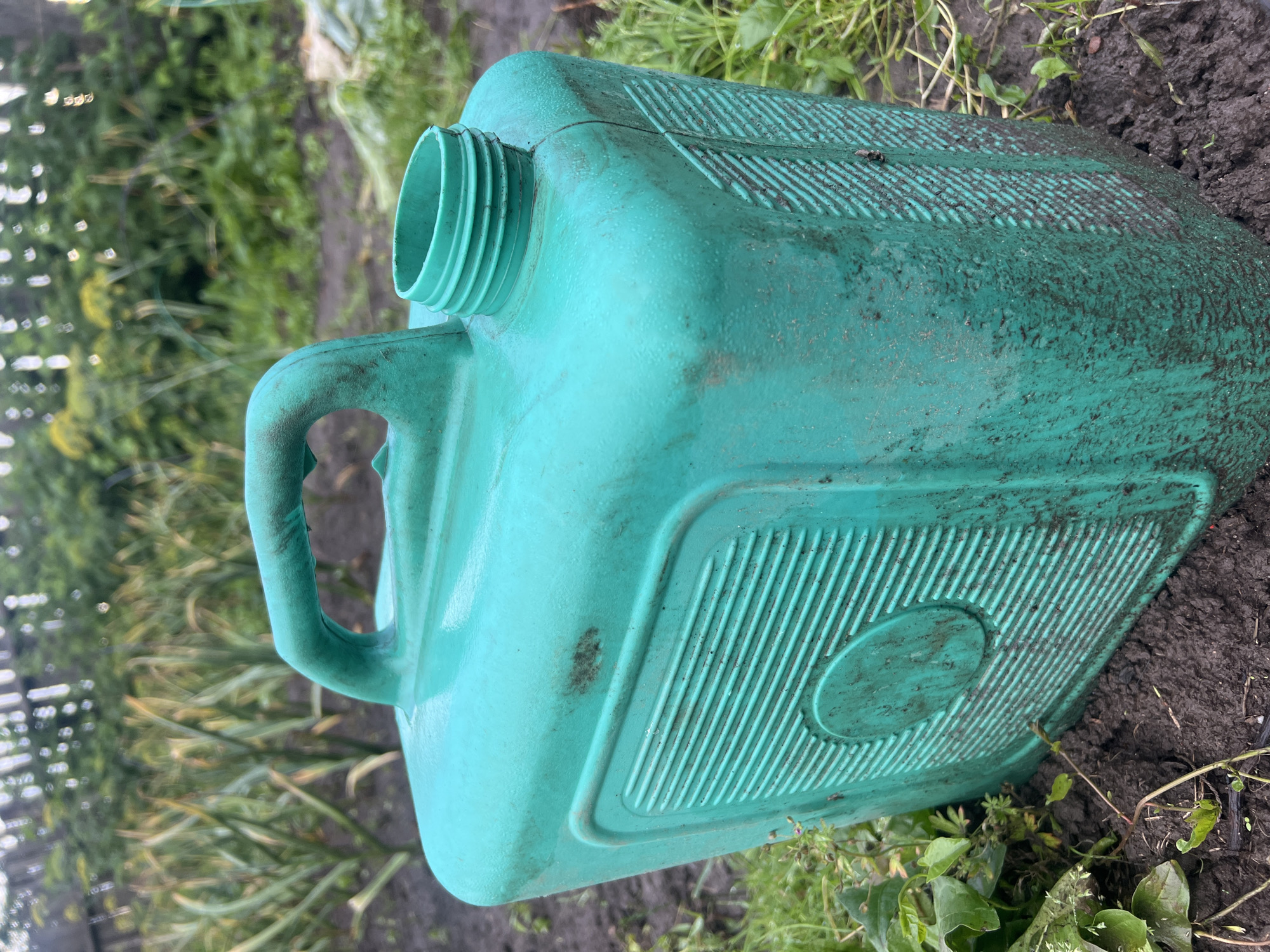 The image shows a dirty, green plastic jerrycan placed on the ground in an outdoor environment, likely a garden or yard. The jerrycan has a handle and an open spout. The ground is covered with soil, grass, and some dried leaves. In the background, there are green plants and a wooden fence. The overall impression is one of a functional item left outdoors in a slightly unkempt setting. The location is identified as Бучацінскі сельскі Савет, Belarus.