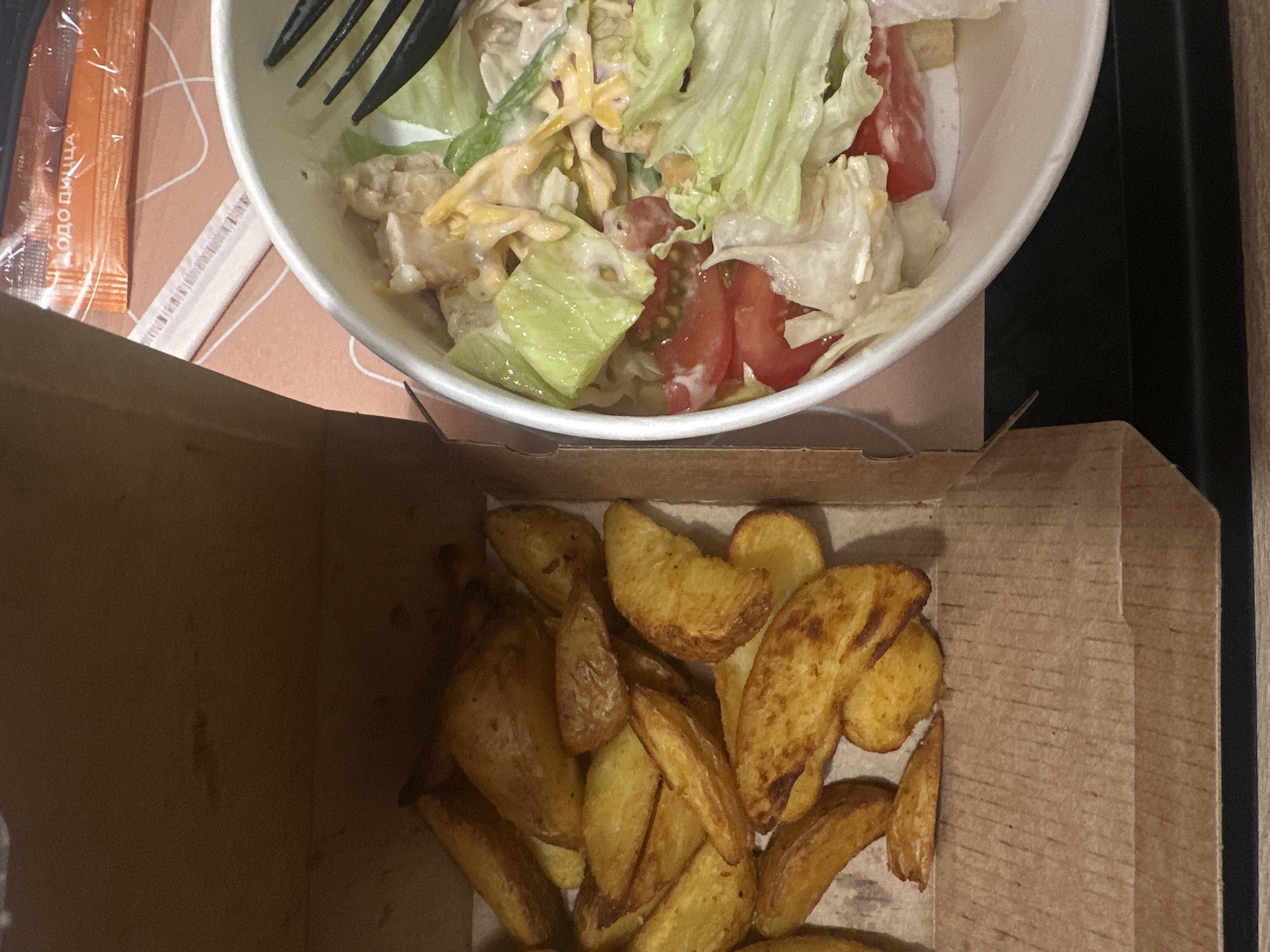 An overhead shot captures a casual meal featuring a salad and potato wedges in takeaway containers.
In the upper part of the frame, a white bowl holds a vibrant salad. It primarily consists of fresh green and lighter-colored lettuce leaves, bright red tomato pieces, and what appears to be shredded chicken or cheese, all coated in a creamy white dressing. A black plastic fork is partially visible, resting within the salad.
To the left of the salad bowl, an orange packet with text and a white, tubular wrapper (possibly for a straw or stirrer) are placed on a patterned, light brown surface. The text clearly visible on the orange packet reads "ДОДО ПИЦЦА" (Dodo Pizza).
Below the salad, a brown cardboard takeaway box is filled with numerous golden-brown potato wedges. These appear to be roasted or fried, with some pieces showing crispier, darker edges.
The scene suggests an indoor setting, possibly a table, where a meal from a takeaway or delivery service is being enjoyed. The lighting is ambient, typical of indoor conditions. There are no people visible in the image. Given the "Dodo Pizza" text, the meal is likely sourced from this Russian chain, aligning with the provided location context of Votkinsk, Russia.