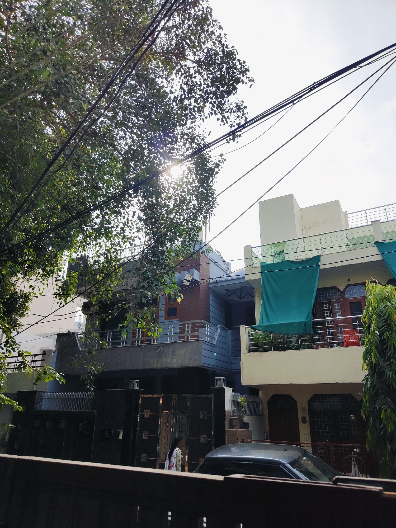 This image depicts a street scene in Gurgaon, India, characterized by residential buildings and overhead electrical wires. In the foreground, a dark-colored car is parked. To the left, a tall, dark gate with intricate metalwork is visible, partially obscured by foliage. Behind the gate, a woman wearing a light-colored patterned garment is walking. The buildings in the background are multi-story structures with balconies. One building features a large, teal-colored tarp suspended over its balcony. Trees with dense green foliage are present, with sunlight filtering through the leaves, creating lens flare in the upper portion of the image. The sky is bright and overcast. The architecture suggests a residential neighborhood in an urban or suburban Indian setting.