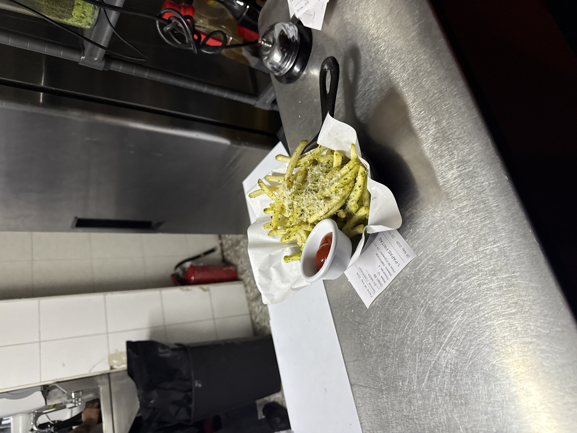 A tilted medium shot shows a small cast iron skillet filled with green fries, possibly pesto fries, with parmesan cheese grated on top, along with a small white ramekin with ketchup. The skillet is placed atop a printed receipt with Spanish writing on it, which reads: "Fecha 08 may 2026 Terminal. (1)SERVER1, EN MESA-Orden: 88 Mesero: ABRAHAM Mesa 12 Personas: 4, Hora de Impresión: 23:13 11, 1-PAPAS FRITAS, 08 May, 2026 23:13". The skillet sits on a stainless steel countertop in what appears to be a restaurant kitchen. The countertop features numerous scratches and abrasions. The kitchen wall and floor are tiled. Behind the skillet are other kitchen equipment and supplies, as well as a small fire extinguisher. Based on the receipt, the scene is located in Mexico City, Mexico.