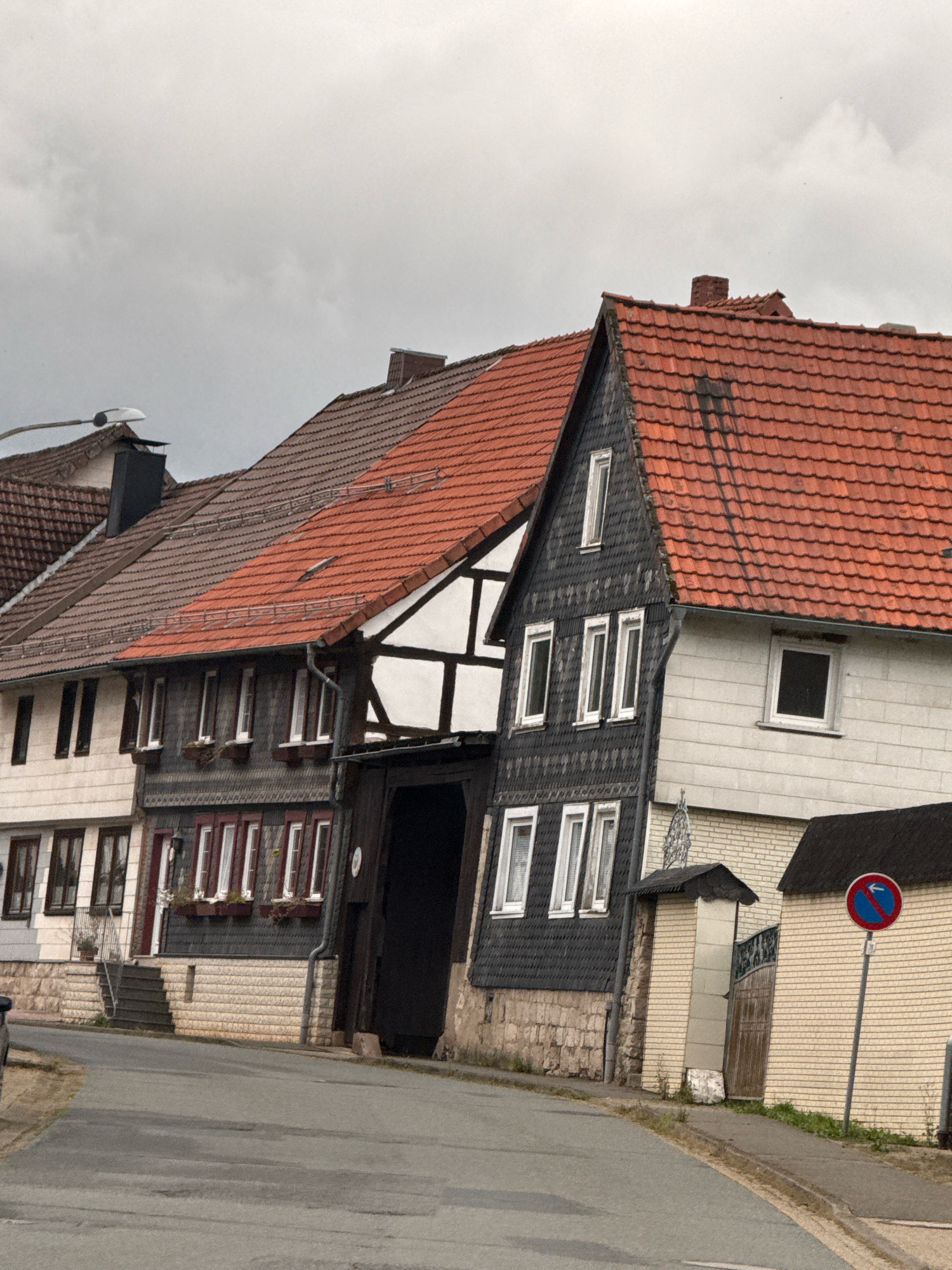The image depicts a street scene in Bad Sachsa, Germany, on a cloudy day. The view is of several traditional German houses with red tiled roofs. The houses are multi-story structures, some featuring dark wooden or slate siding and white trim around windows. The street itself is paved and appears to be curving. In the foreground, a portion of a car is visible on the left. On the right side of the street, a yellow brick building stands next to a gate, and a "no parking" sign with a blue circle and a red diagonal line, indicating a parking prohibition, is posted on a pole. The sky is overcast, suggesting the possibility of rain or a generally gloomy weather.