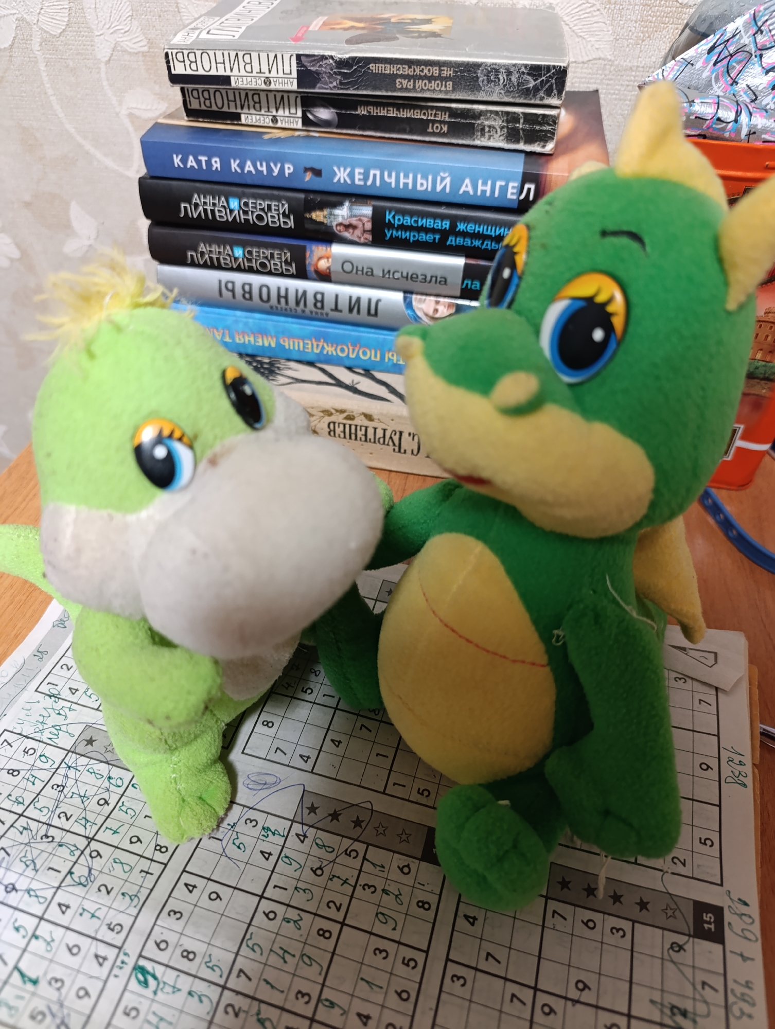 A medium shot shows two green plush toy dragons sitting on a piece of paper with a sudoku puzzle filled in with pencil marks. Behind the toys is a stack of books. The books include a book by Katya Kachur, two books by Anna and Sergei Litvinov, and others whose titles are partially obscured. The background is a light-colored wall. To the right is part of an orange container.