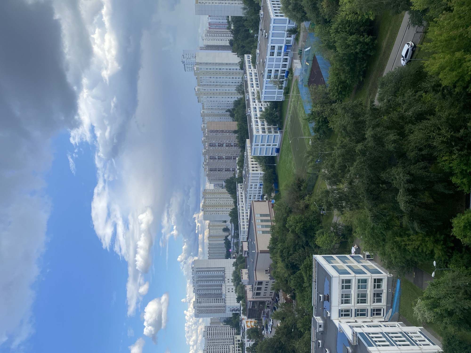 The image captures a residential area in Khimki, Russia, under a partially cloudy sky. Dominating the scene are numerous multi-story apartment buildings, varying in height and architectural style, with many featuring light-colored facades. In the foreground and middle ground, several lower-rise buildings, possibly educational institutions or community centers, are visible. These buildings are predominantly white with blue accents and large windows. Lush green trees and vegetation surround the buildings, interspersed with paved areas, sidewalks, and a road. On the road, a white car is parked. Recreational facilities, including a sports field with a track and a fenced court with playground equipment, are also present. The sky transitions from bright blue with scattered white clouds on the left to darker, more dramatic cloud formations on the right, suggesting a changing weather pattern. The overall impression is of a developed urban environment integrated with significant green spaces.