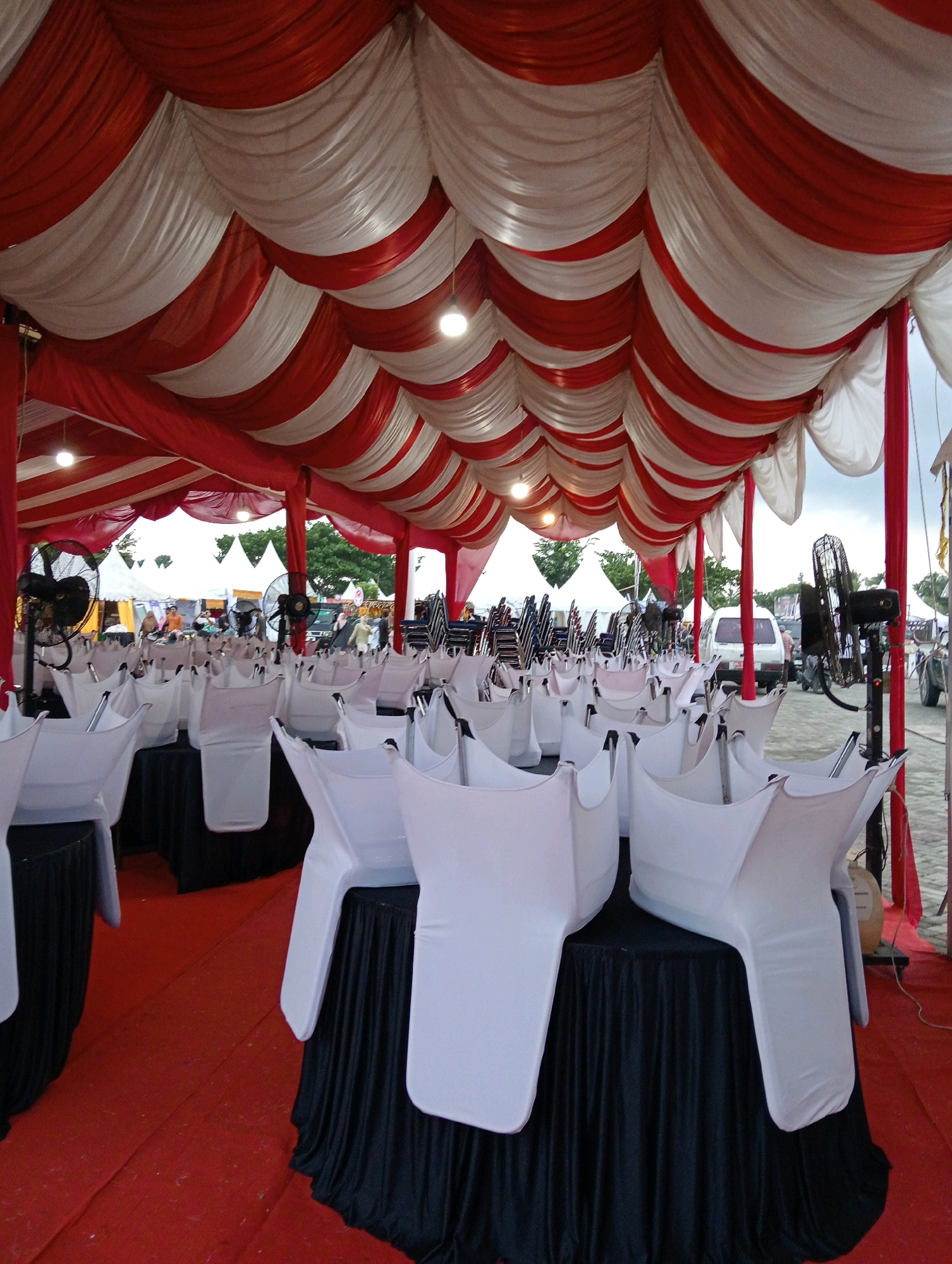 The image depicts an outdoor event setup under a red and white striped tent. Rows of white-covered chairs are placed around tables draped with black cloths. Fans are visible, positioned to provide air circulation. In the background, several white tents are set up, suggesting a larger event. A white van and other vehicles are also present, along with trees and a cloudy sky, indicating an outdoor location. The red carpeted floor suggests a formal or festive occasion. The scene appears to be in Meunasahaluedrienlhoksukon Barat, Indonesia, likely a public area prepared for an event.